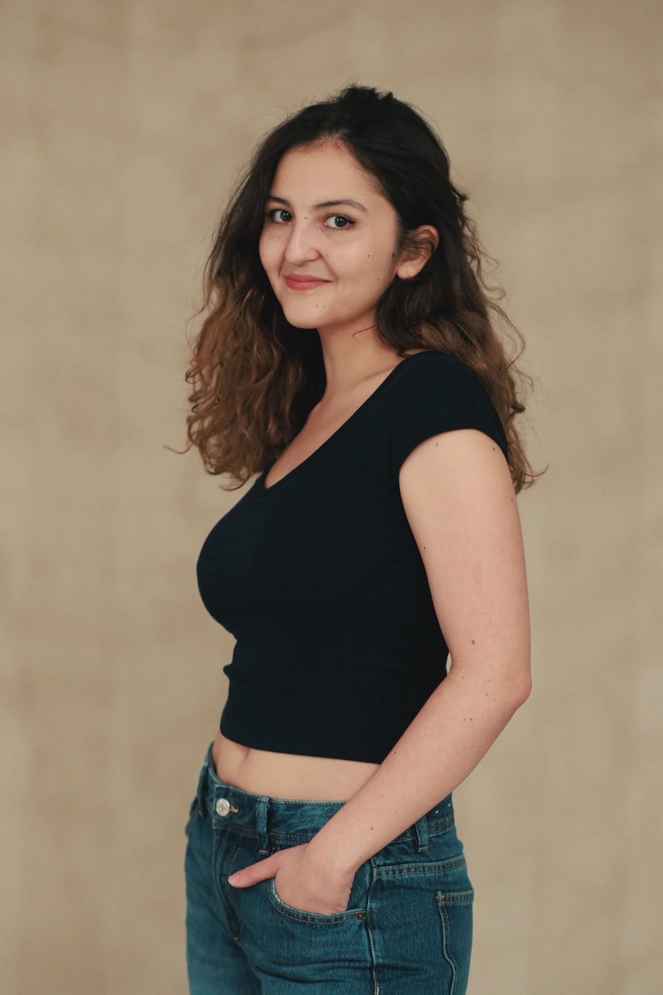 Young woman with curly hair wearing a black crop top and jeans, standing against a beige background, looking at the camera with a slight smile.