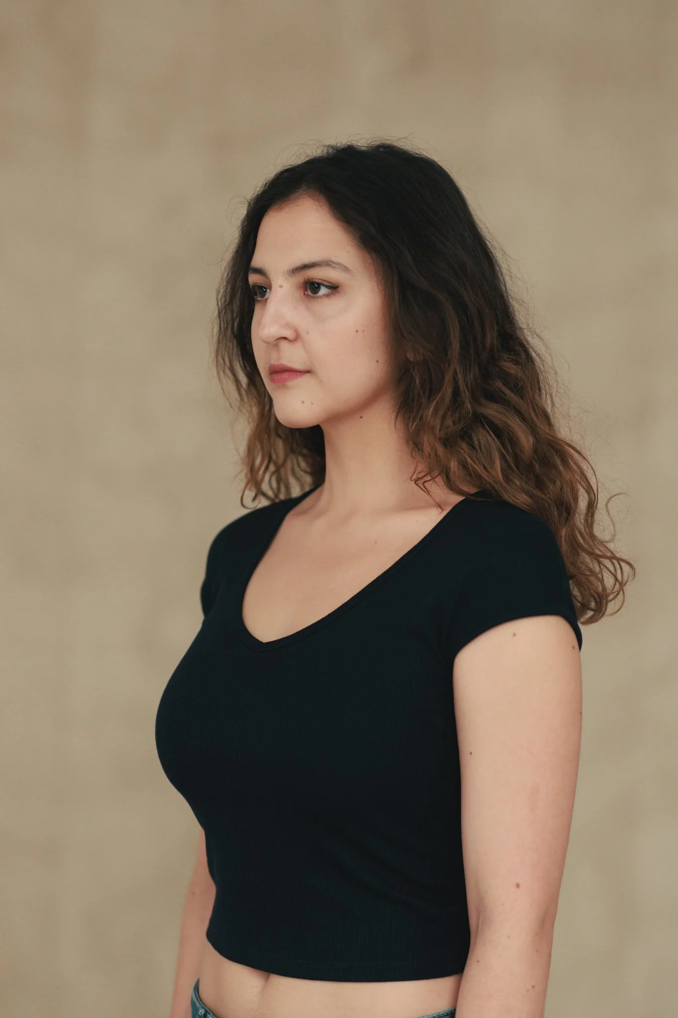 A young woman with shoulder-length wavy brown hair, wearing a black V-neck T-shirt, standing against a neutral background.