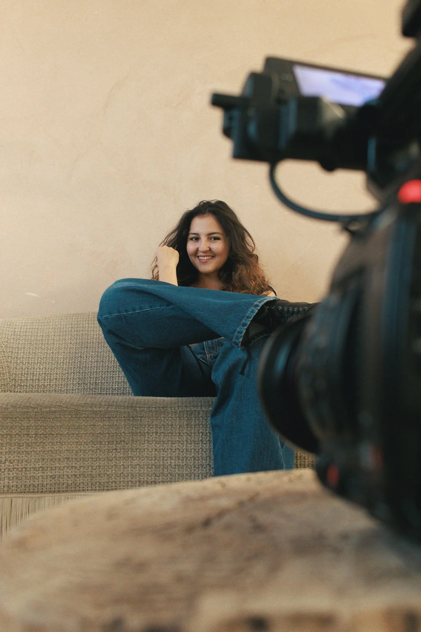 Young woman sitting on a sofa with casual attire, smiling for the camera during a video recording.