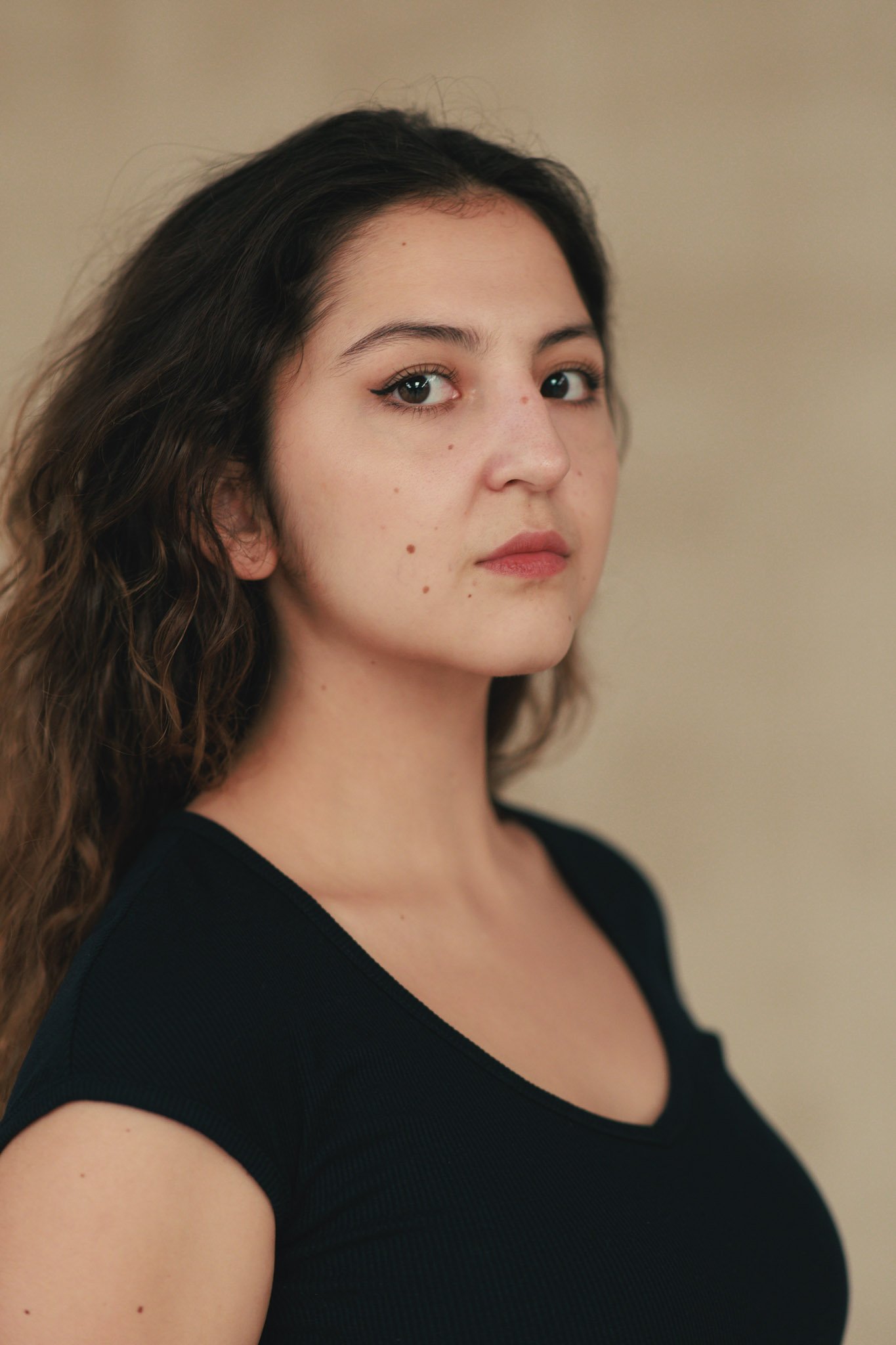 A young woman with dark wavy hair and fair skin looks at the camera with a neutral expression. She is wearing a black top against a plain beige background.