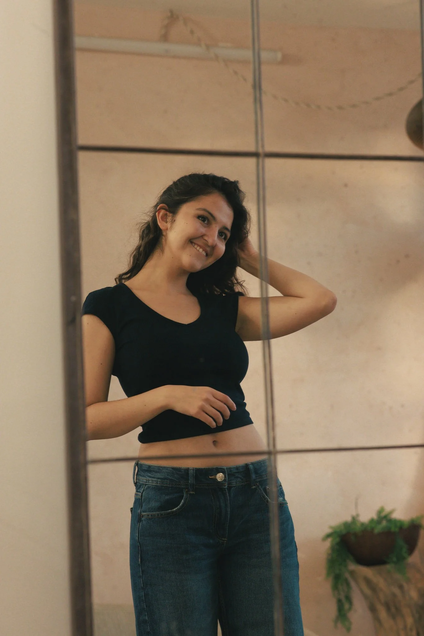 A woman taking a mirror selfie, smiling, with dark curly hair, wearing a black crop top and blue jeans in a room with beige walls and a small potted plant.
