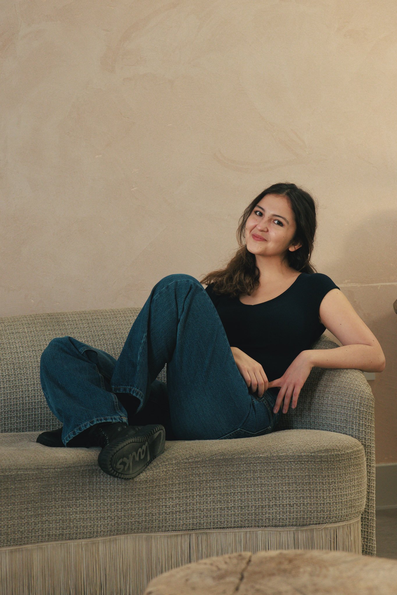 Young woman with long dark hair sitting relaxed on a beige couch, wearing a black t-shirt, blue jeans, and black shoes, smiling at the camera.