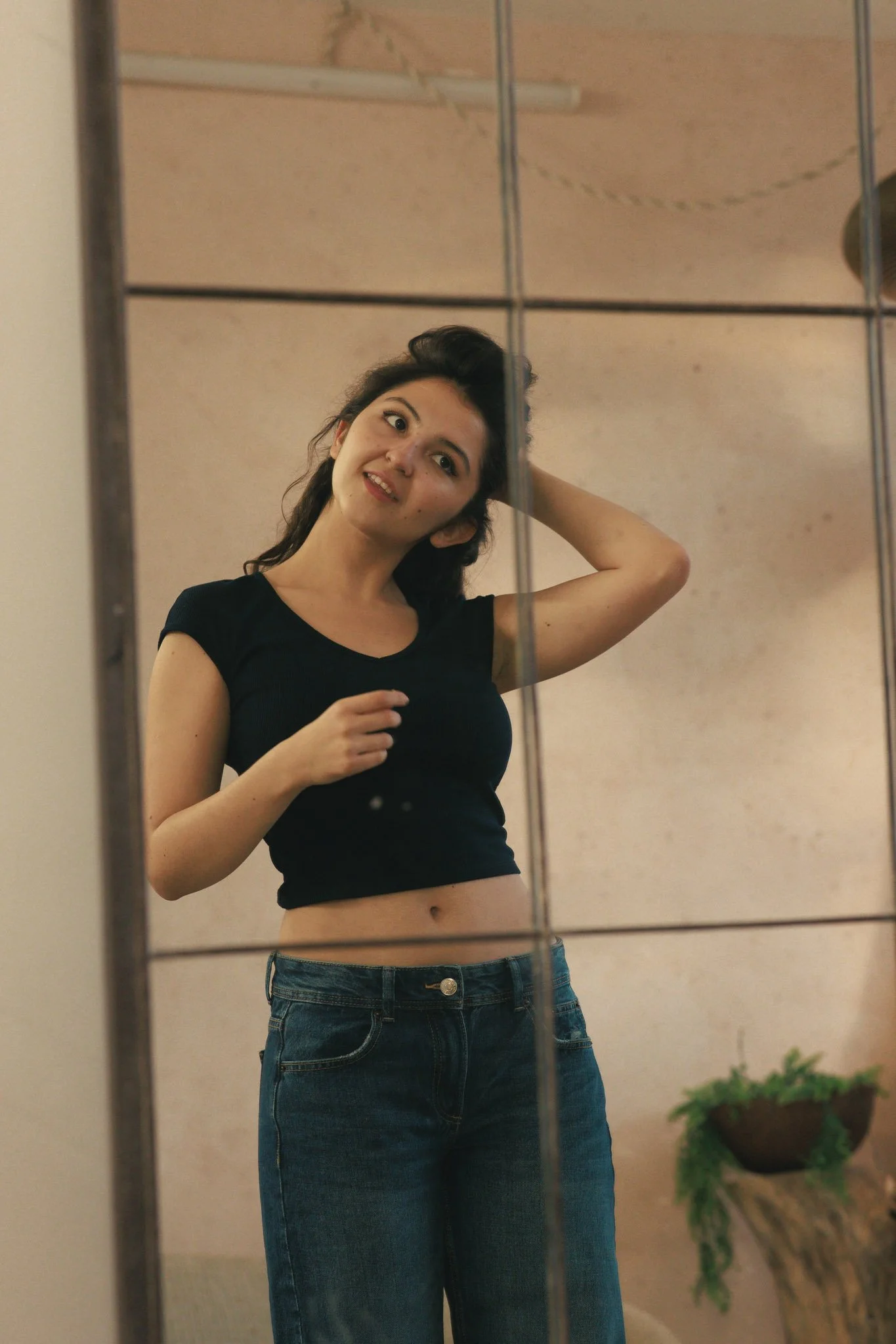 Young woman taking a mirror selfie, wearing a black crop top and blue jeans, with a casual indoor background.