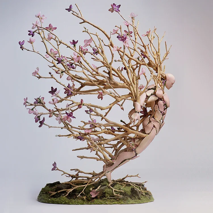 A sculpture of a human figure intertwined with blooming pink and purple flowering branches on a mossy base.