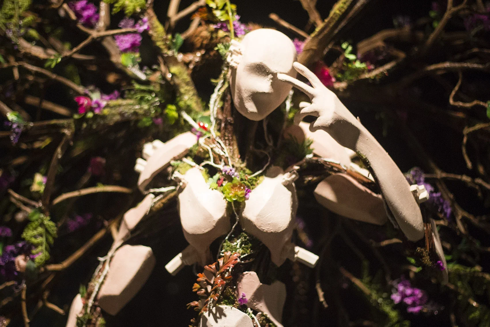 Sculpture of a woman with flowers and greenery around her, posing with her hand near her face, surrounded by dark background with purple flowers.