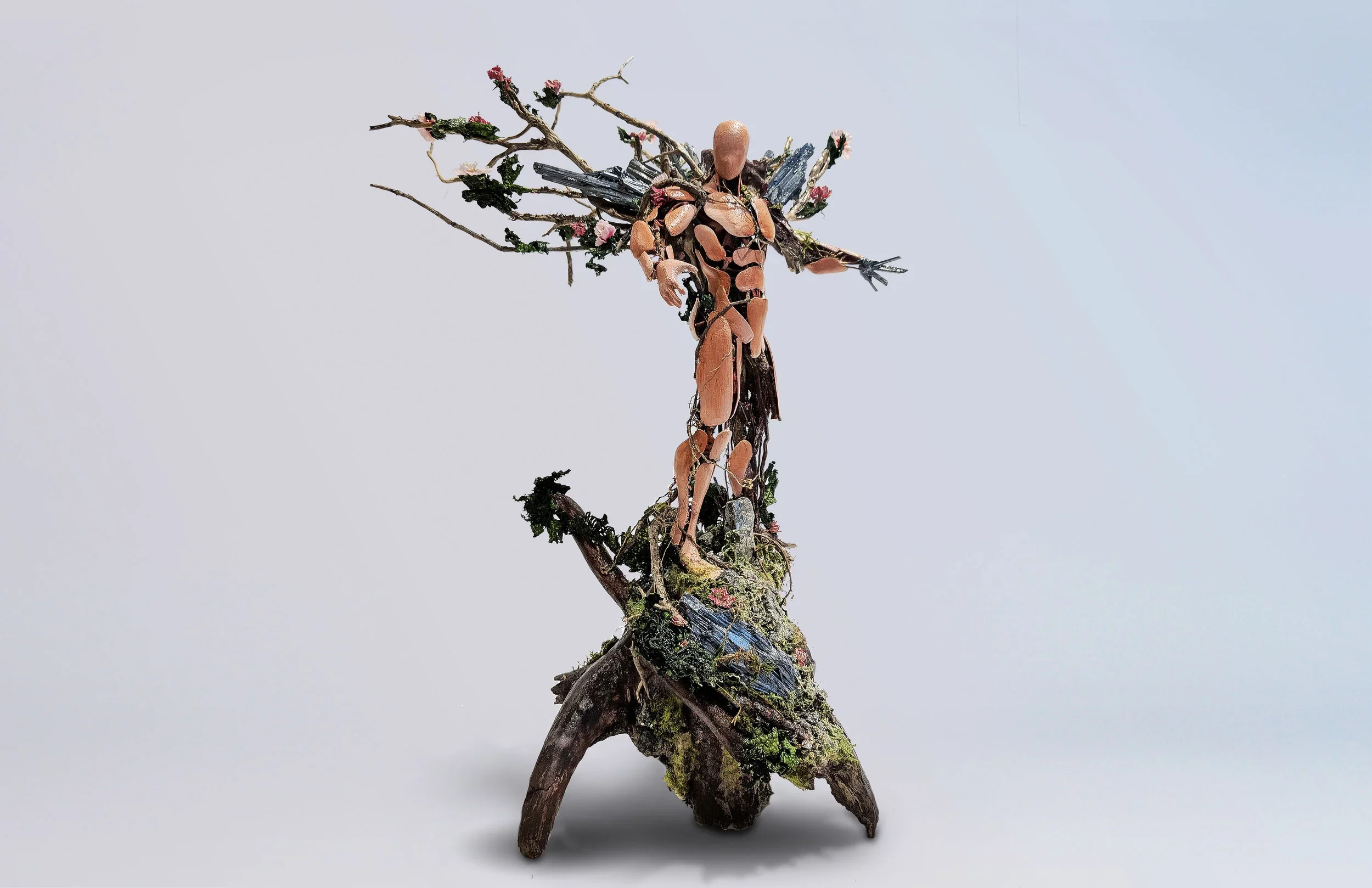 A sculpture resembling Jesus on the cross, made from natural materials including branches, wood, moss, and small flowers, set against a plain background.