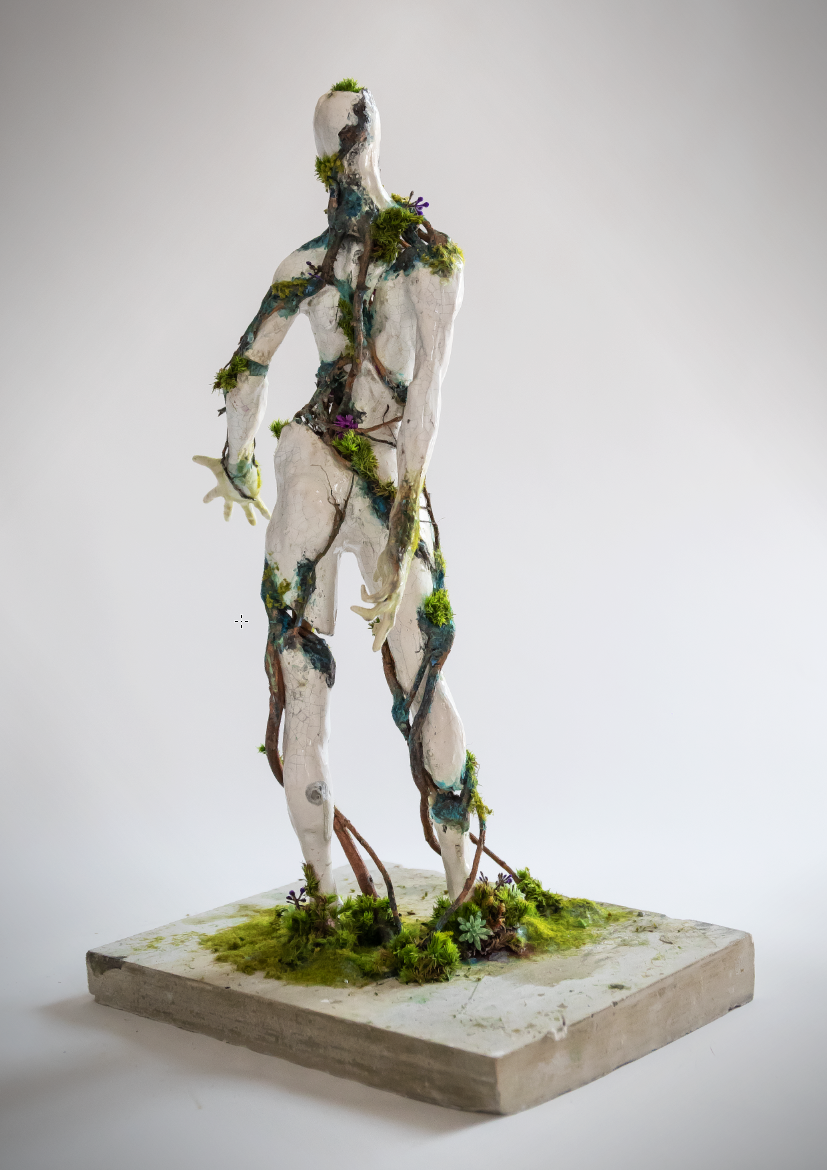A sculpture of a human figure covered in moss, small plants, and vines, standing on a rectangular base with grass and plants. The sculpture appears weathered and cracked, with natural elements integrated into its form.