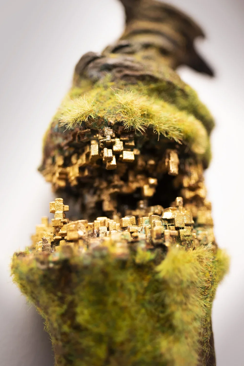 Close-up of a moss-covered, metallic object resembling a tree trunk with tiny, gold-colored blocks and green moss growing on it.