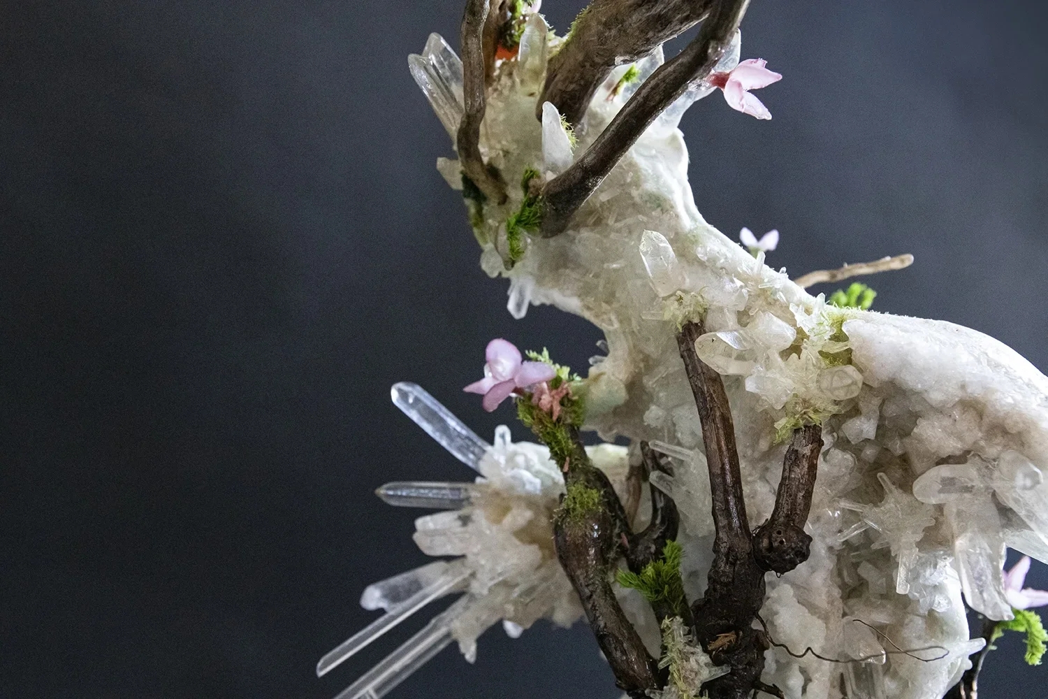A close-up of an art sculpture featuring a mix of natural elements like twigs, small flowers, and crystalline structures, set against a dark background.