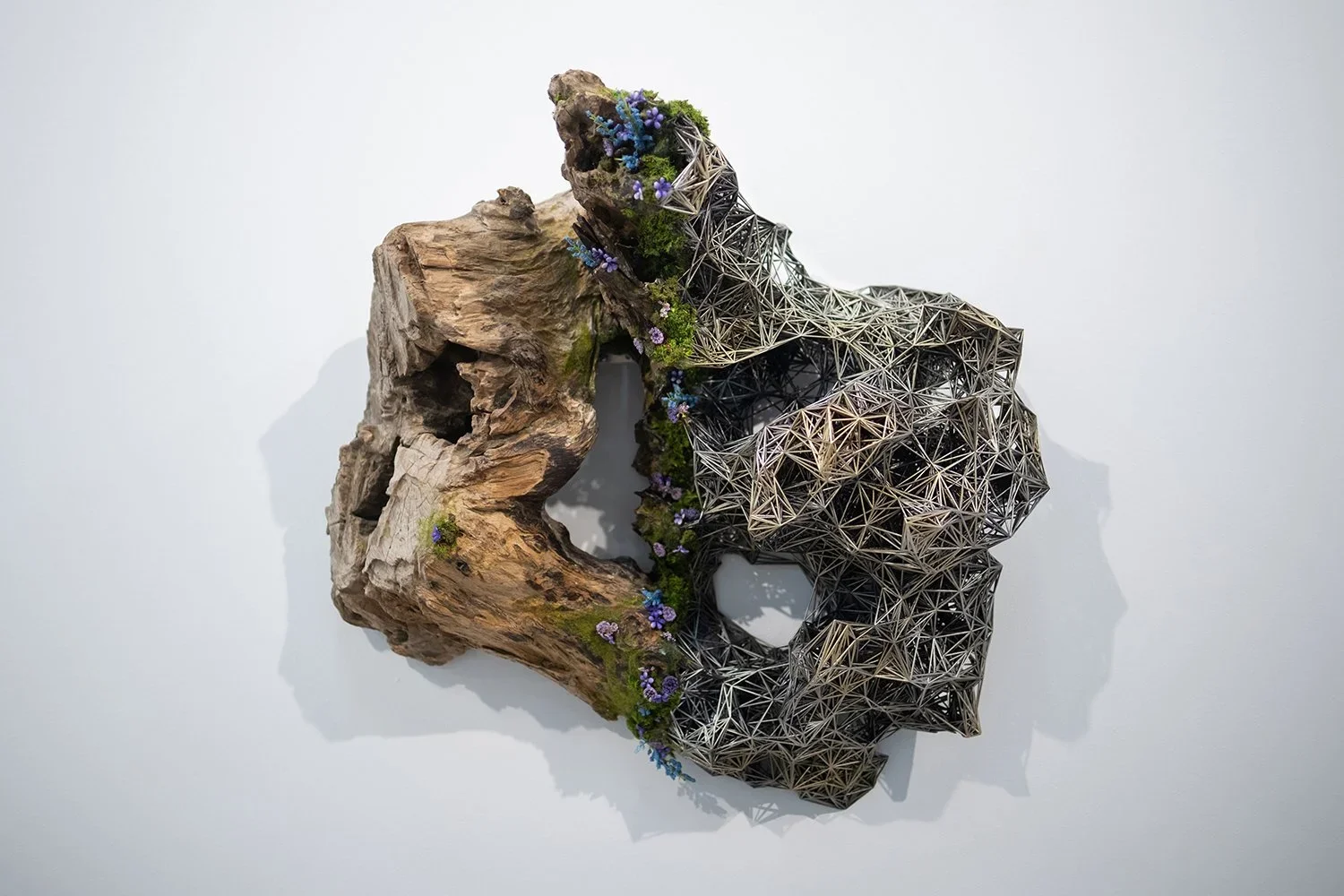 Mixed media art piece combining a large piece of driftwood with geometric metal sculpture and purple flowers and greenery.