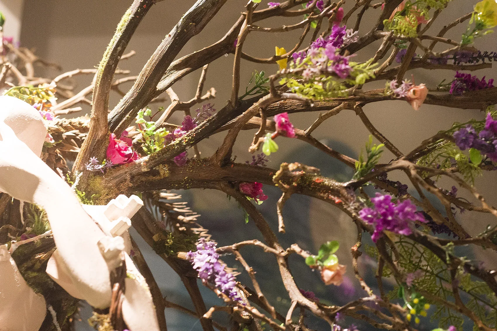 Close-up of a decorative floral arrangement with intertwined brown branches and small pink, purple, and yellow flowers, partially wrapped by a light pink ribbon.