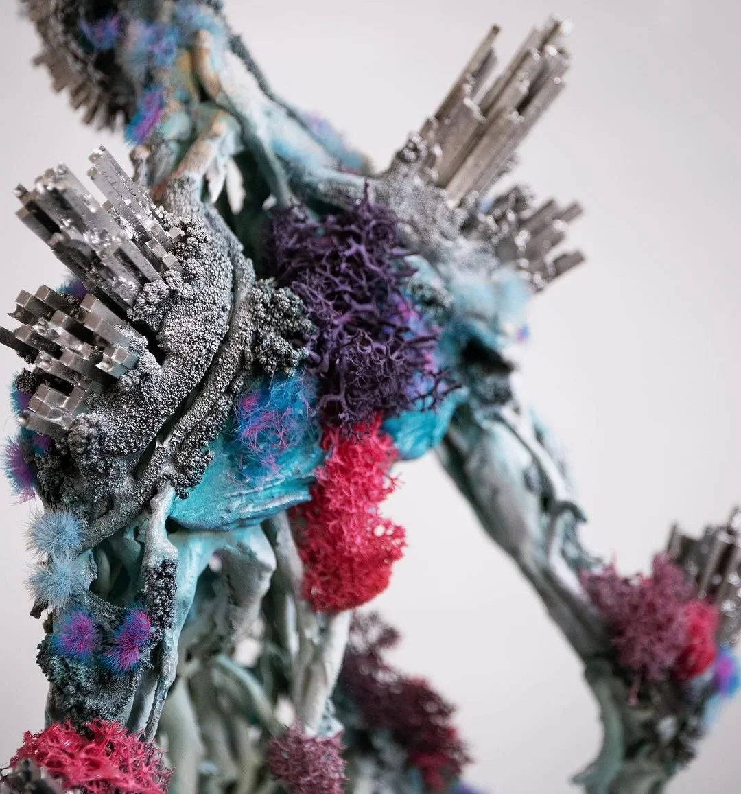 A mixed media sculpture of a humanoid figure made from metallic and textured materials, decorated with colorful fibers and powders.