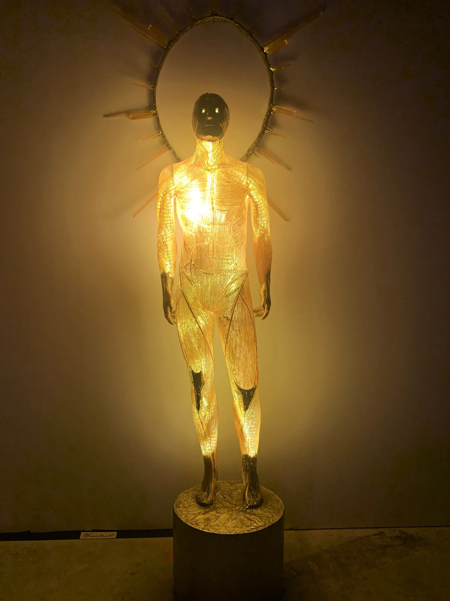 A sculpture of a human figure with a glowing body, standing on a round base. The figure has a halo-like ornament around its head with rays extending outward. The sculpture appears to be made of a translucent, glowing material, with intricate details 