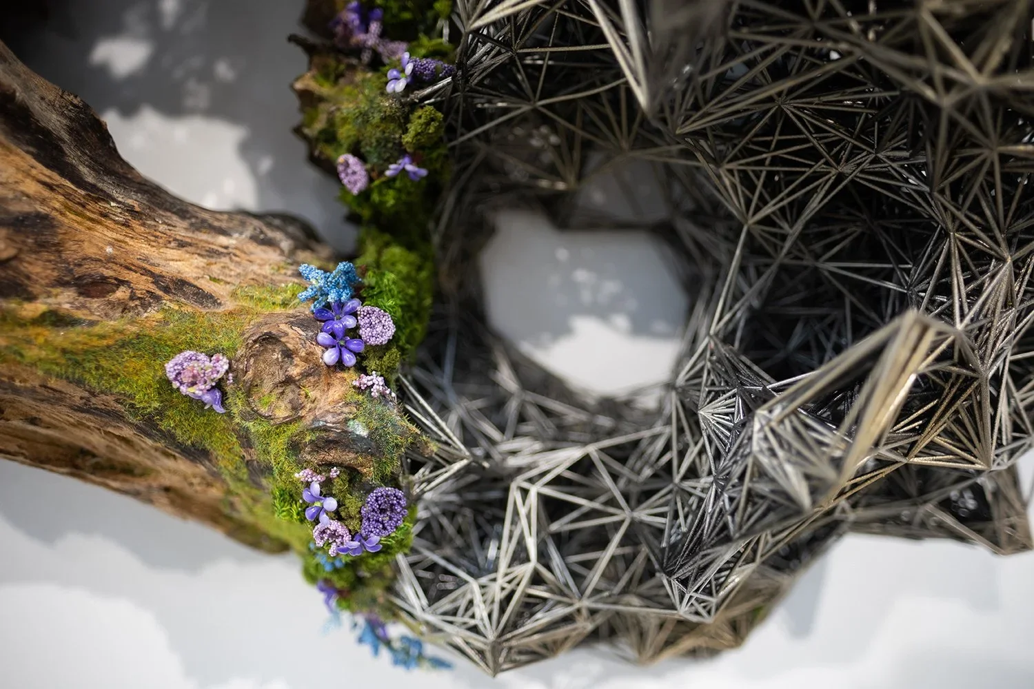 Close-up of a decorative art piece combining a mossy, textured log with tiny purple and blue flowers on one side, and a geometric, metal wire frame with a hollow center on the other side.