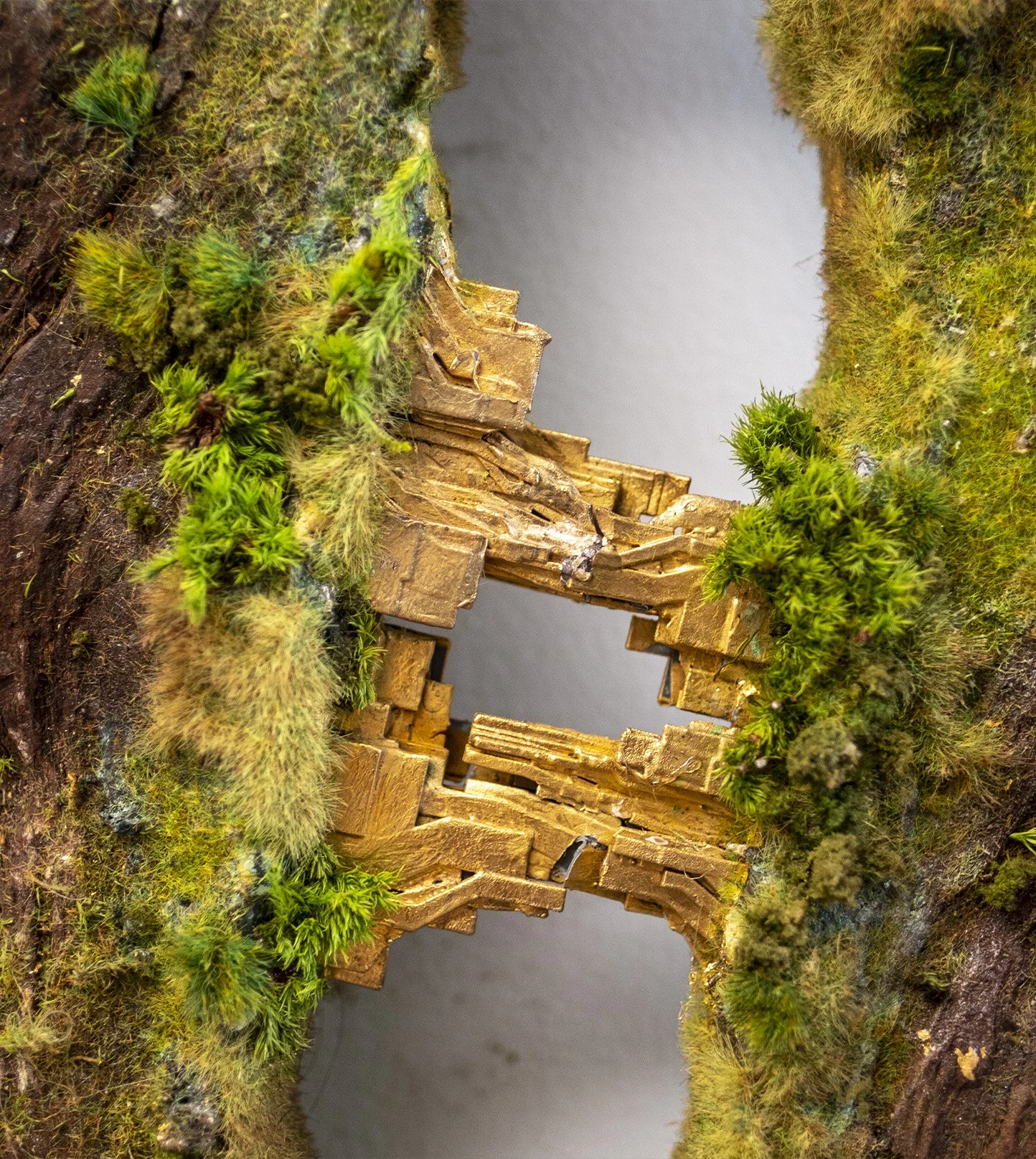 Close-up of a miniature landscape with a small castle or structure made of gold-colored material, nestled among green moss and grass, with a background of a rocky surface.