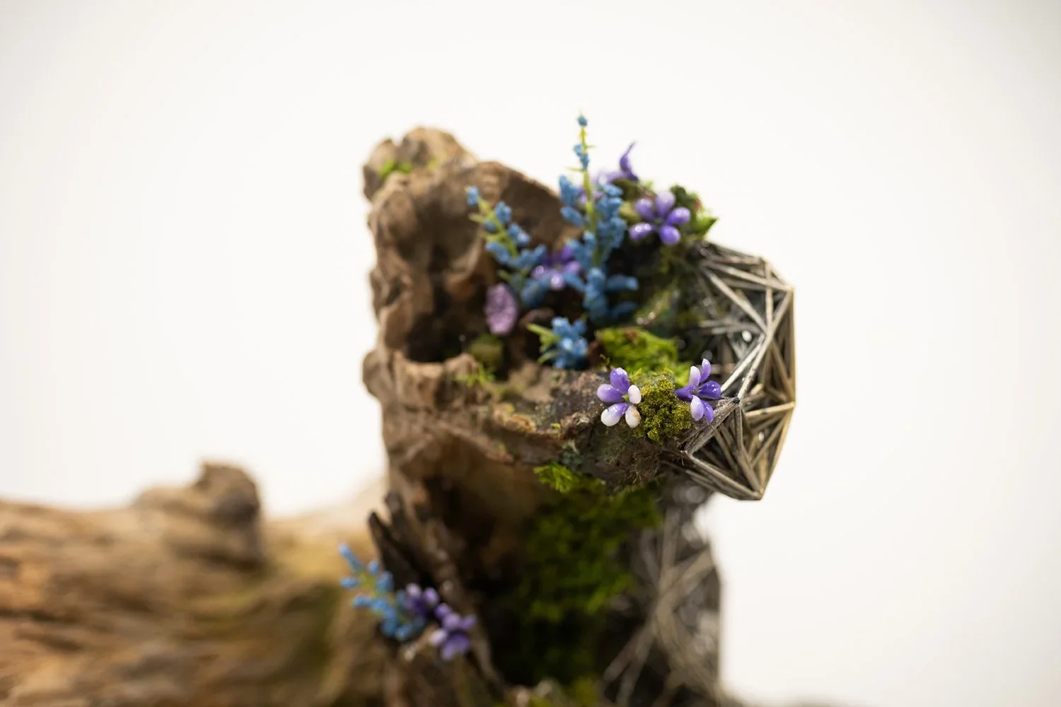 A close-up of a small decorative arrangement featuring dried wood, purple and blue tiny flowers, green moss, and a geometric metallic cone, set against a white background.