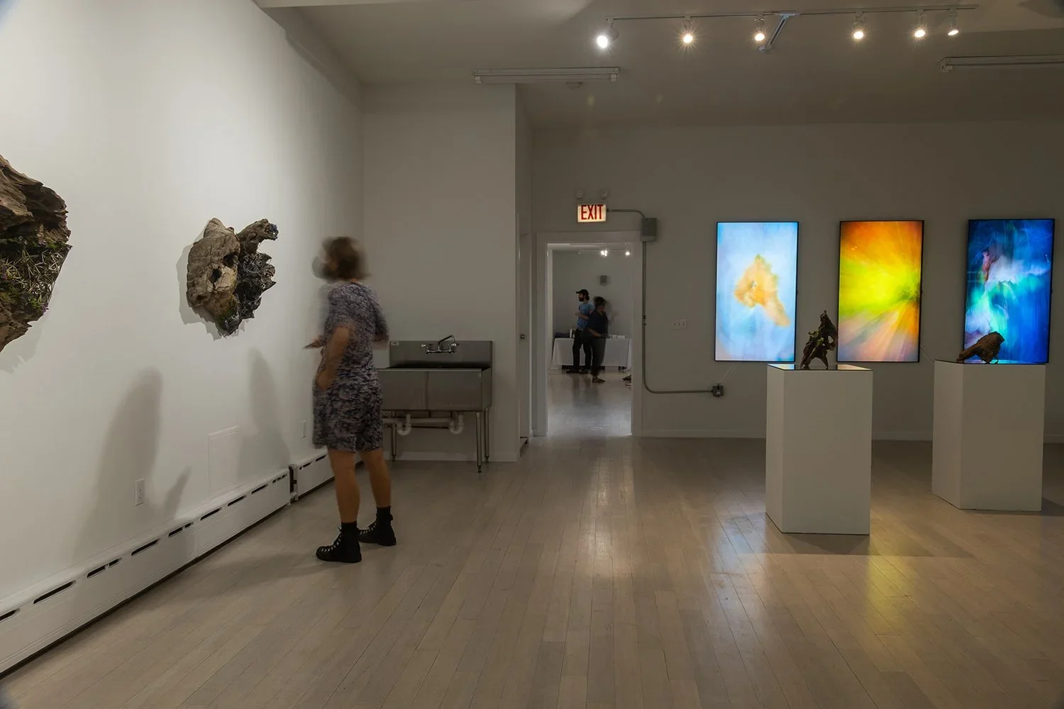 People viewing art exhibits in a gallery, with three vibrant digital artworks on the wall and sculptures displayed on pedestals, seen through an open door.