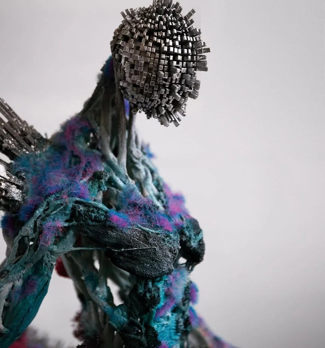 A sculpture of a human figure made from various metal objects, including screws and wire, with colorful fabric or yarn blended into the body and a detailed, textured head.