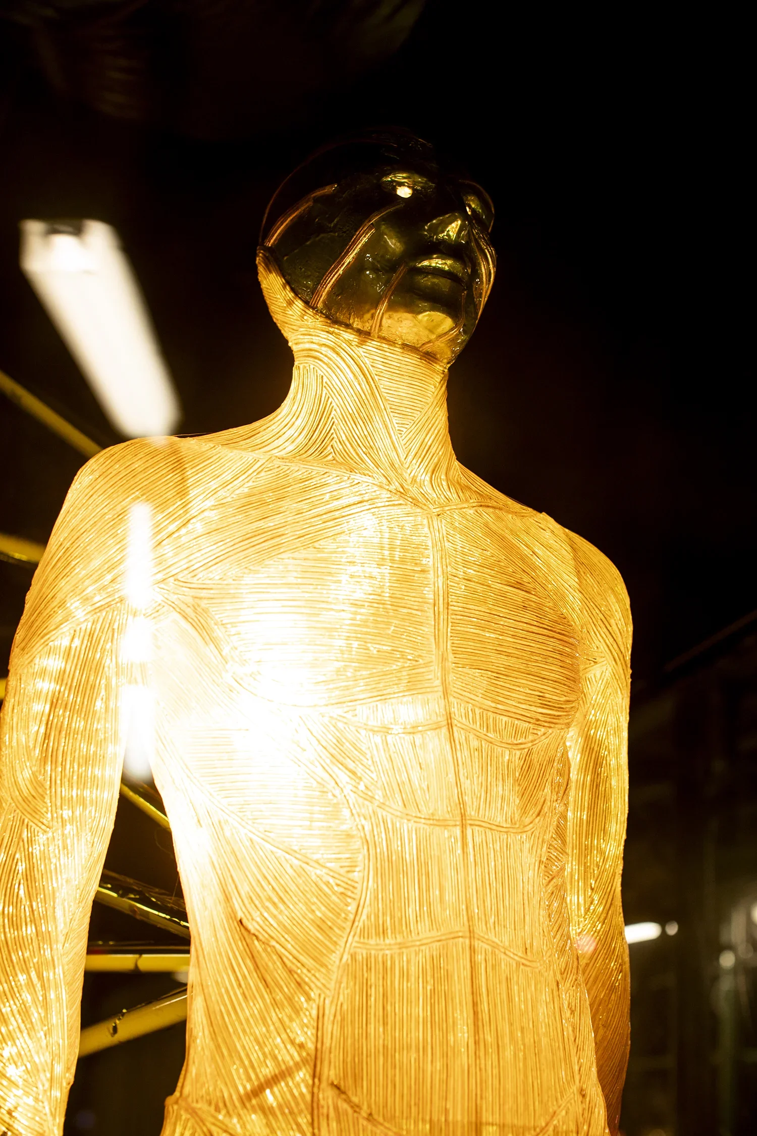 A glowing sculpture of a human figure with a translucent head and detailed body lines, illuminated from within, against a dark background.