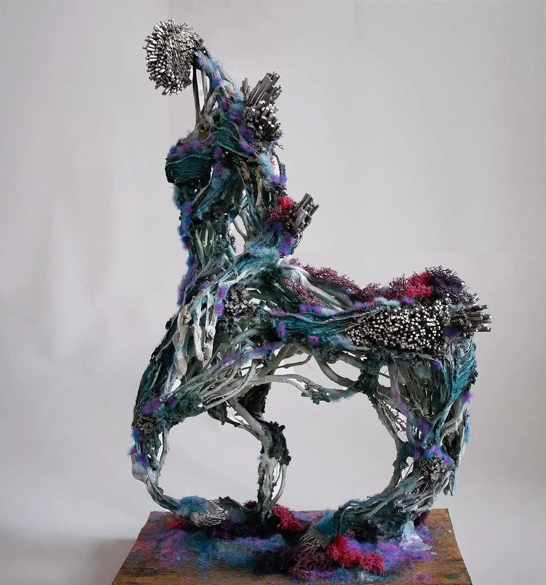 Mixed media sculpture resembling a human figure sitting on a chair, made of metal, fabric, and colorful synthetic materials.