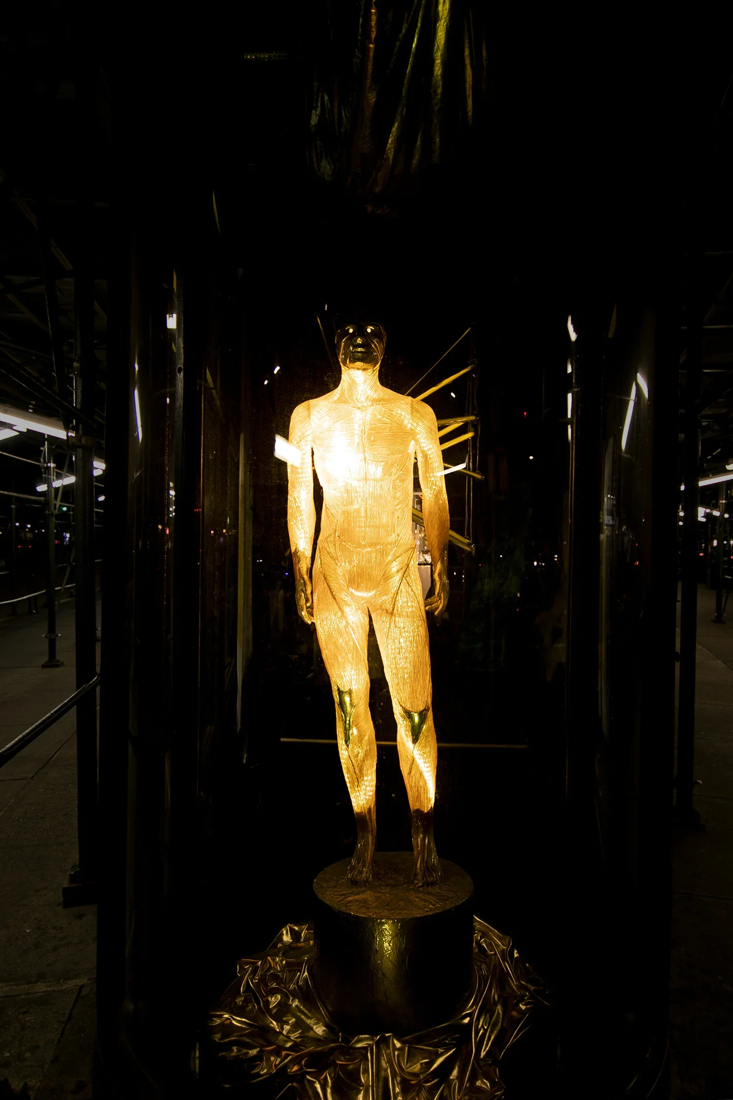 Golden sculpture of a standing female figure illuminated and displayed indoors, with dark surroundings and reflective surfaces.