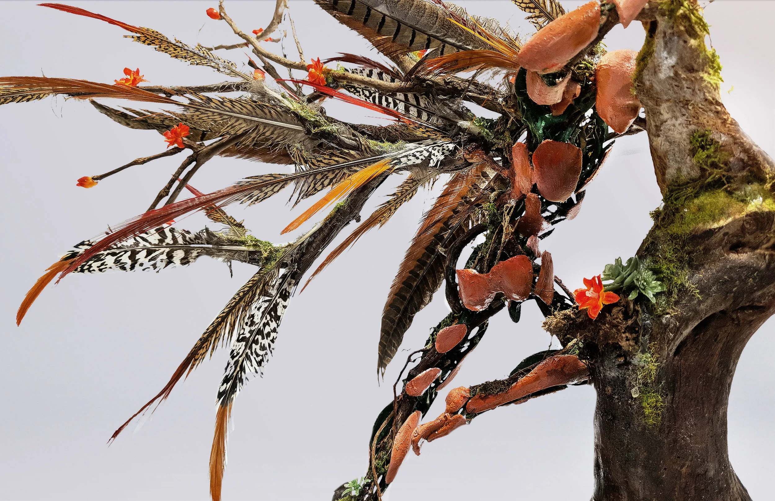 Close-up view of a decorative art piece with feathers, small orange flowers, moss, and clay elements attached to a tree limb.