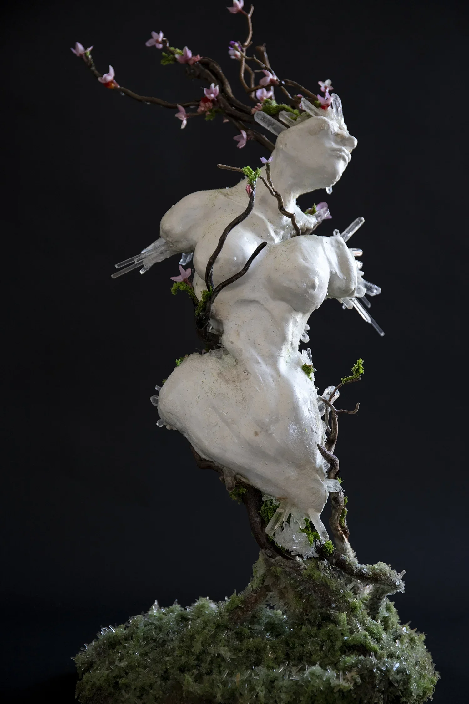 A sculpture of a human face with branches and pink flowers growing from the top and sides, sitting atop a textured white base with green moss and crystal-like formations.