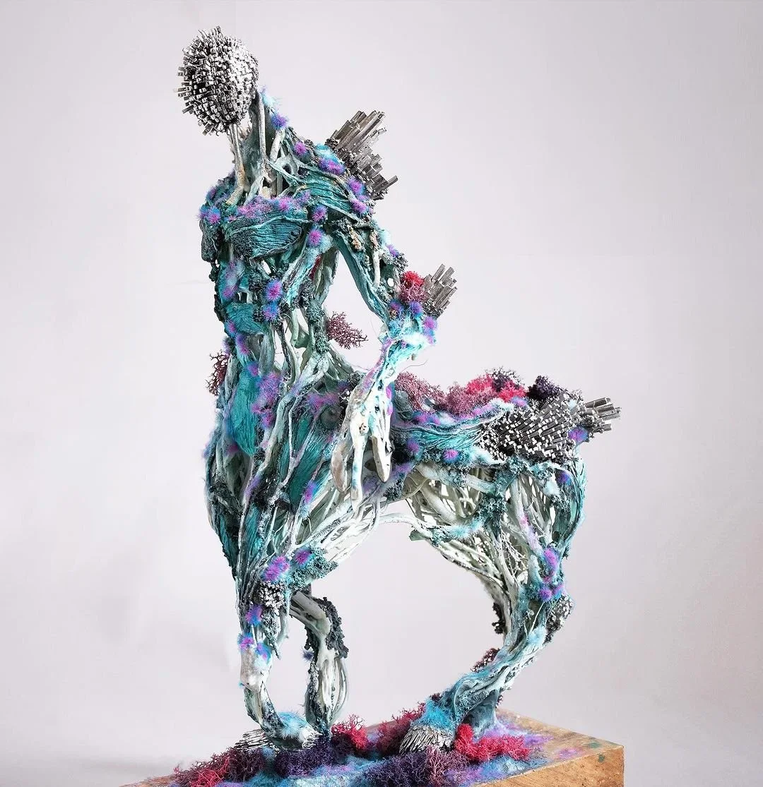 A contemporary sculpture of a humanoid figure with a horse's head, made from mixed media including metal, fabric, and synthetic materials in teal, purple, pink, and black colors.