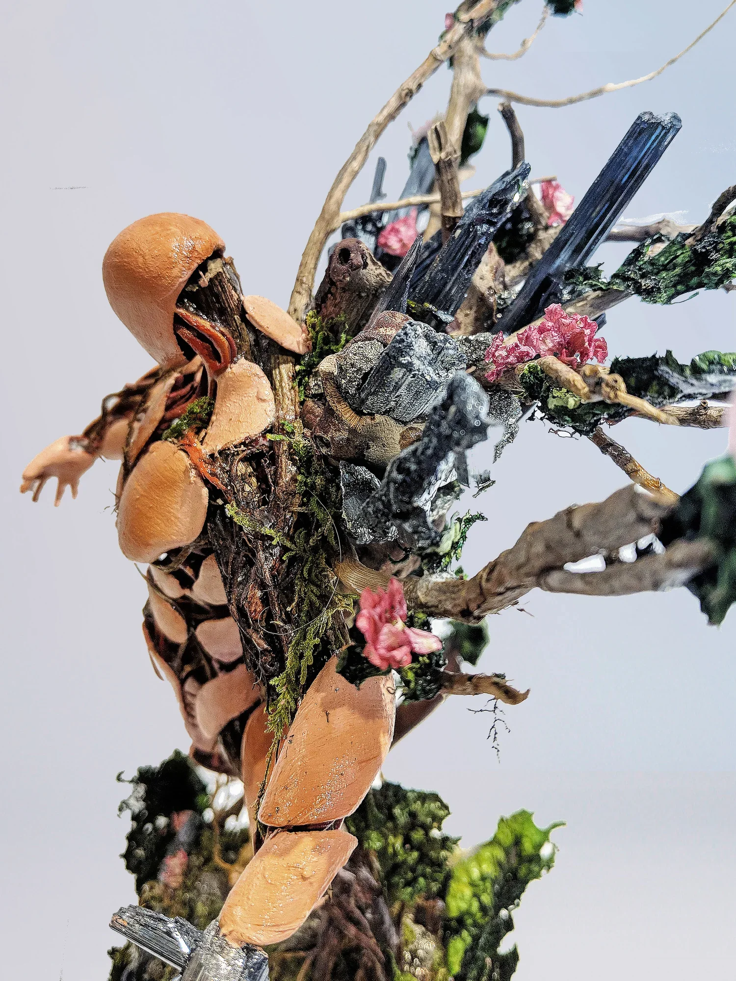 An abstract sculpture made of natural and man-made materials, including wood, metal, moss, and small pink flowers, resembling a creature or plant.