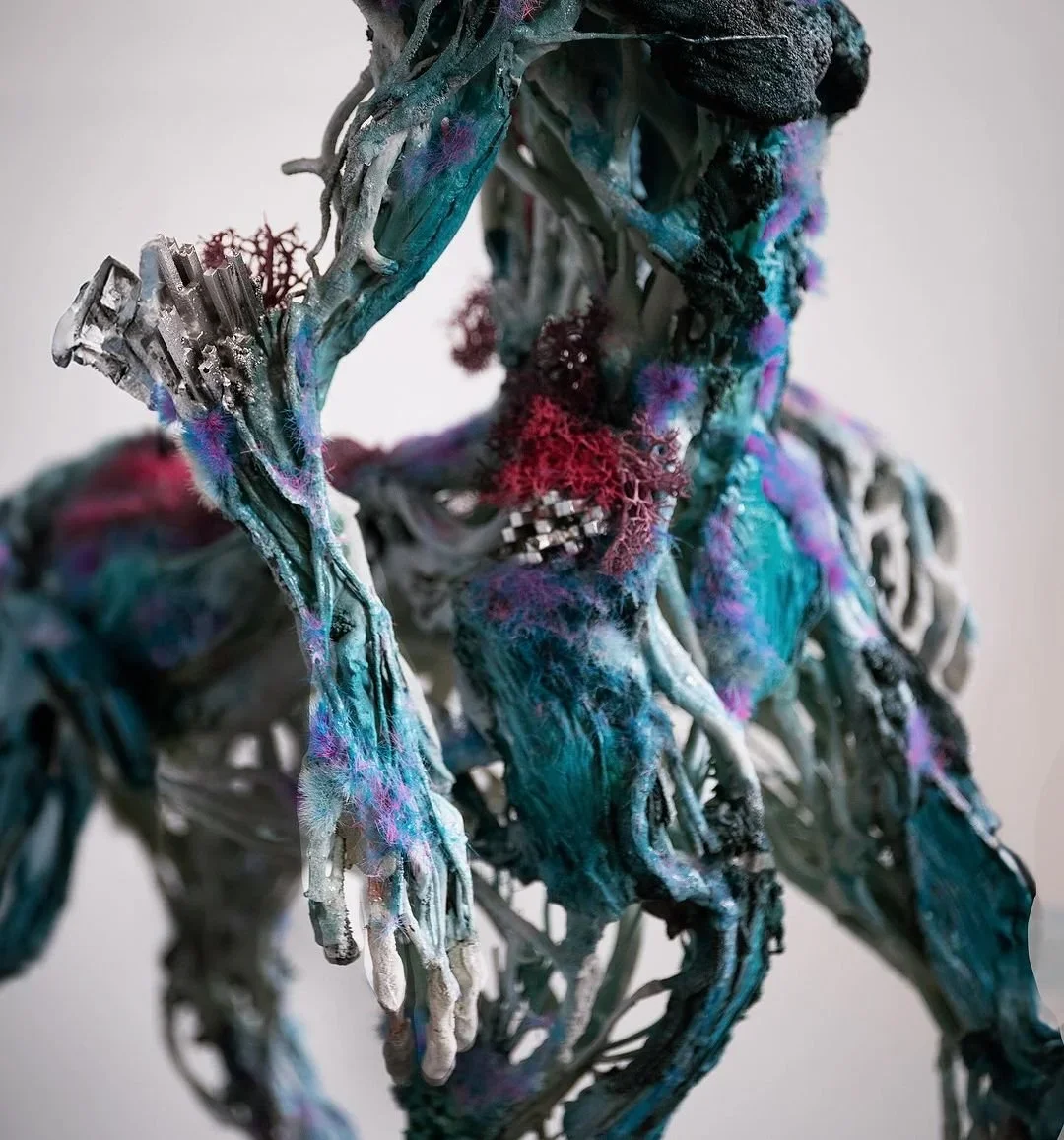 A mixed-media sculpture of a humanoid figure composed of various materials including metal, fabric, and textured elements, with an abstract and industrial appearance.