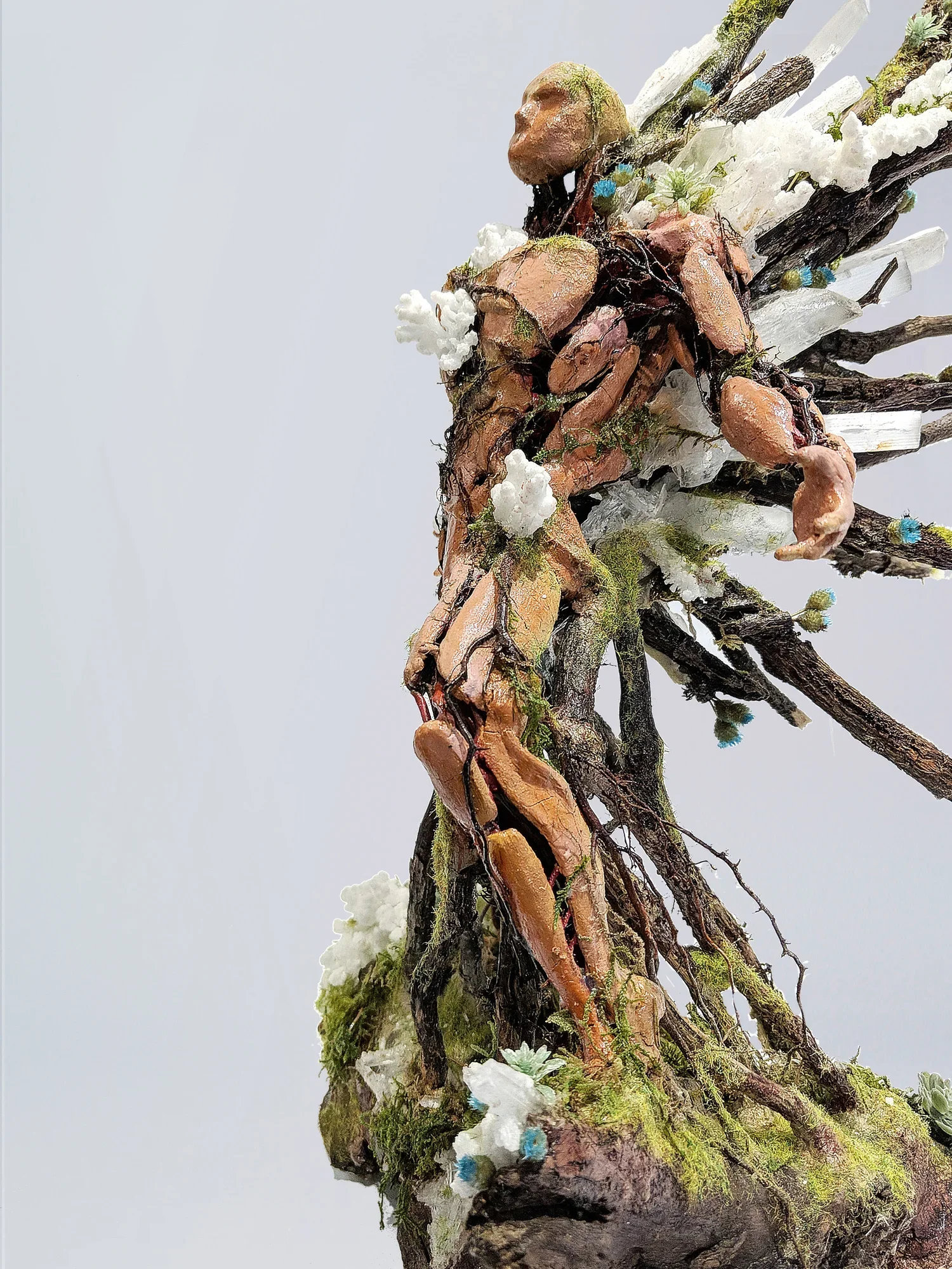A mixed-media art sculpture resembling a tree trunk with branches, adorned with small sculptures, moss, and decorative elements, against a plain white background.