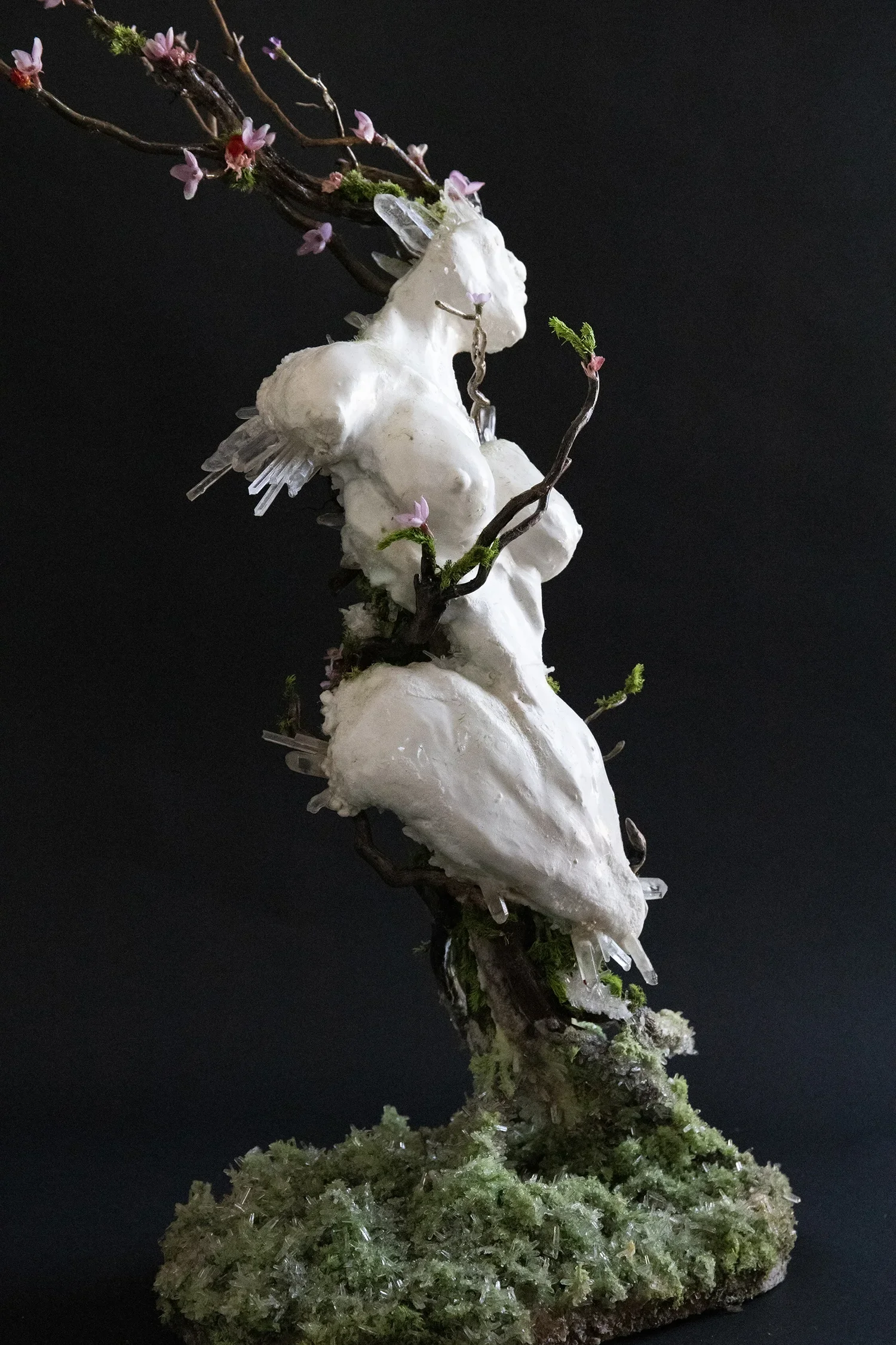 A handcrafted sculpture of a unicorn with a white body, clear ice-like horns and wings, floral decorations, and moss on a textured base against a black background.