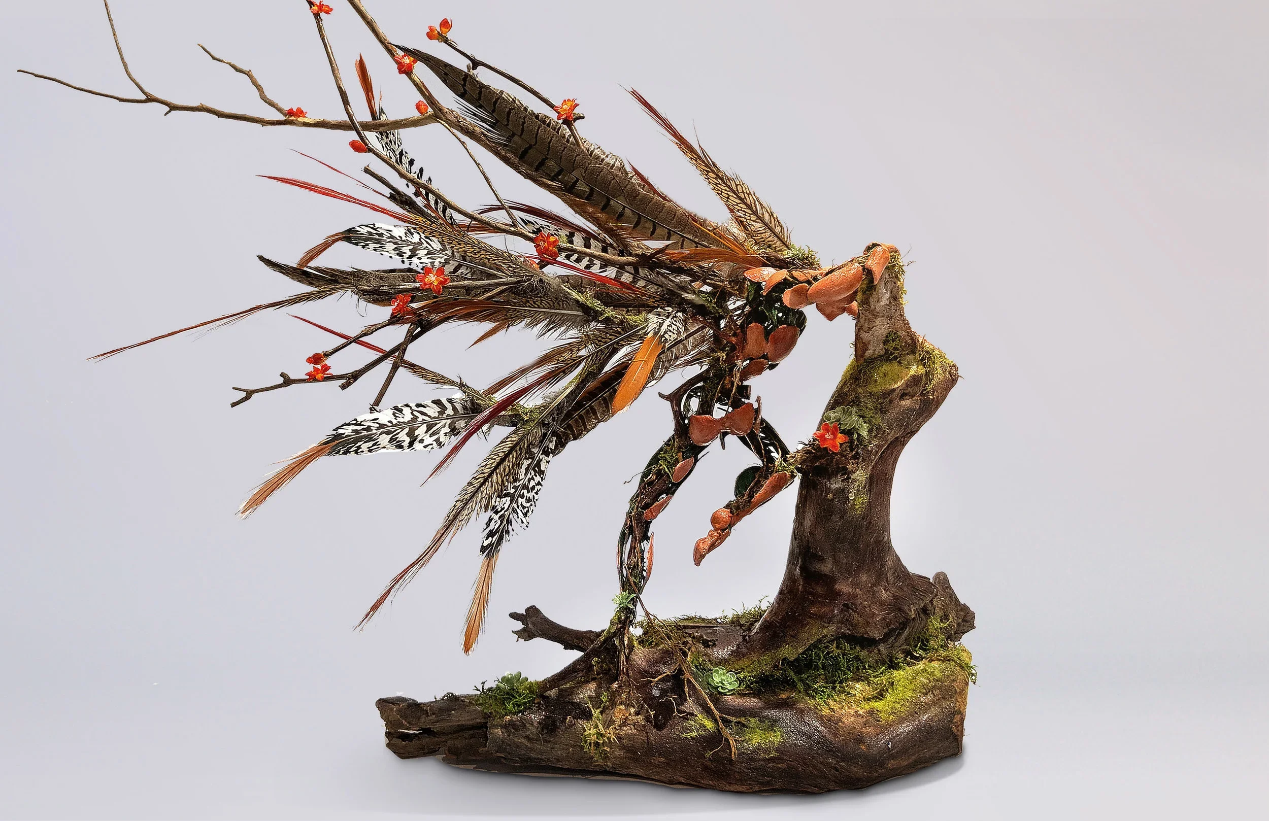 A miniature bonsai tree with a branch-like structure, decorated with colorful feathers and small red flowers, planted in a rustic wooden pot.