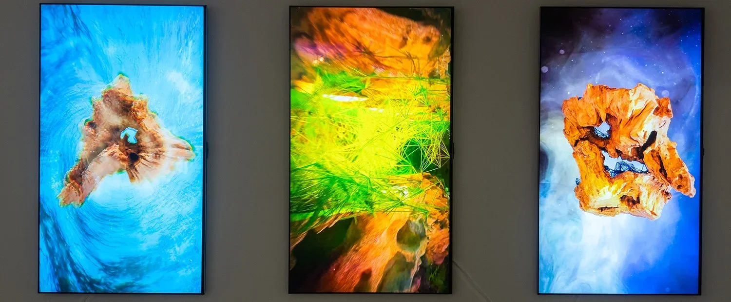 Three digital displays showing abstract artwork. The left screen features a swirling blue and brown pattern, the middle screen displays bright yellow and green interconnected lines and shapes, and the right screen shows a mix of blue, purple, and ora