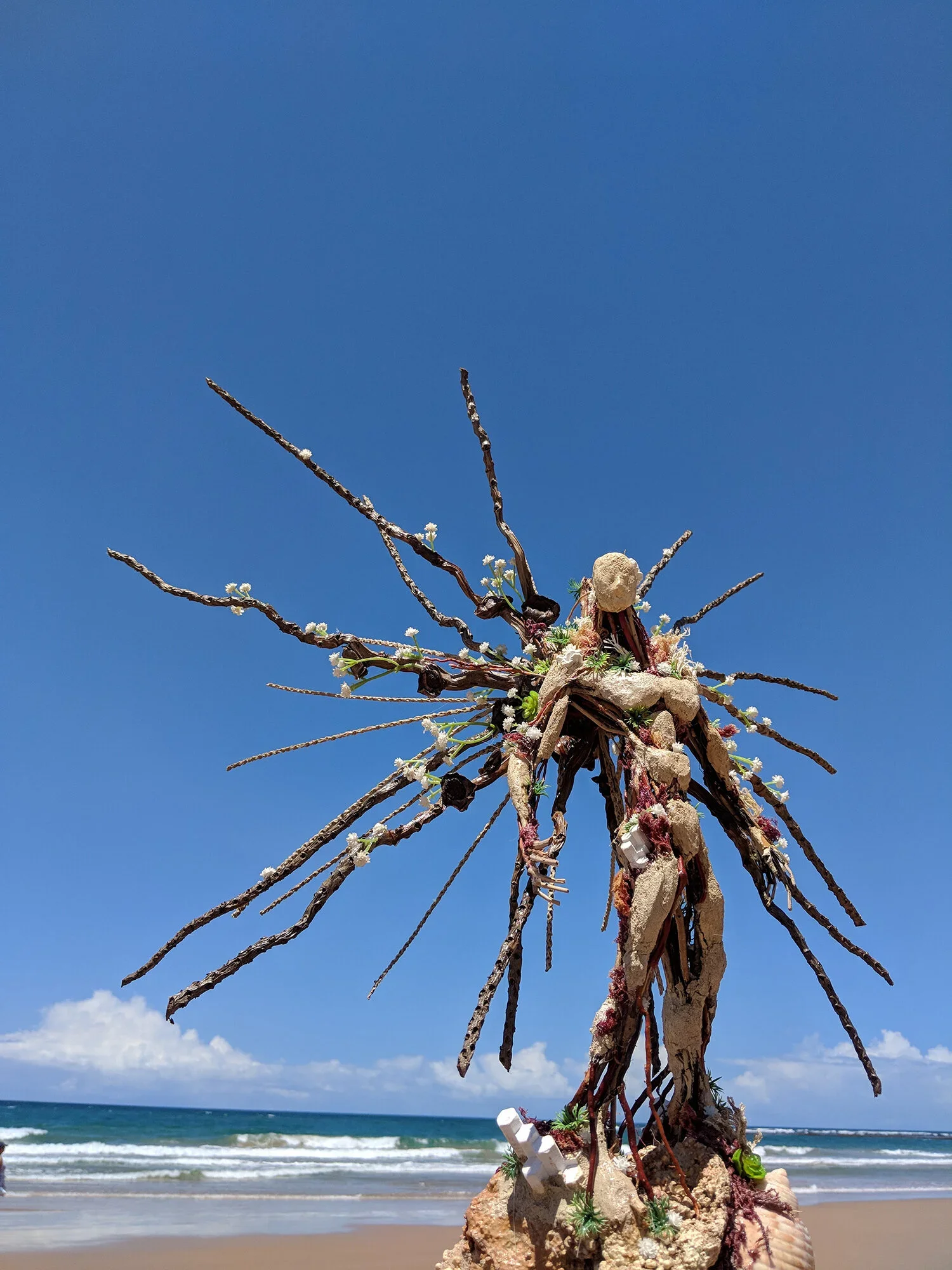 An artistic sculpture resembling a tree made of driftwood, shells, and small plants on a sandy beach with ocean waves and a partly cloudy sky in the background.