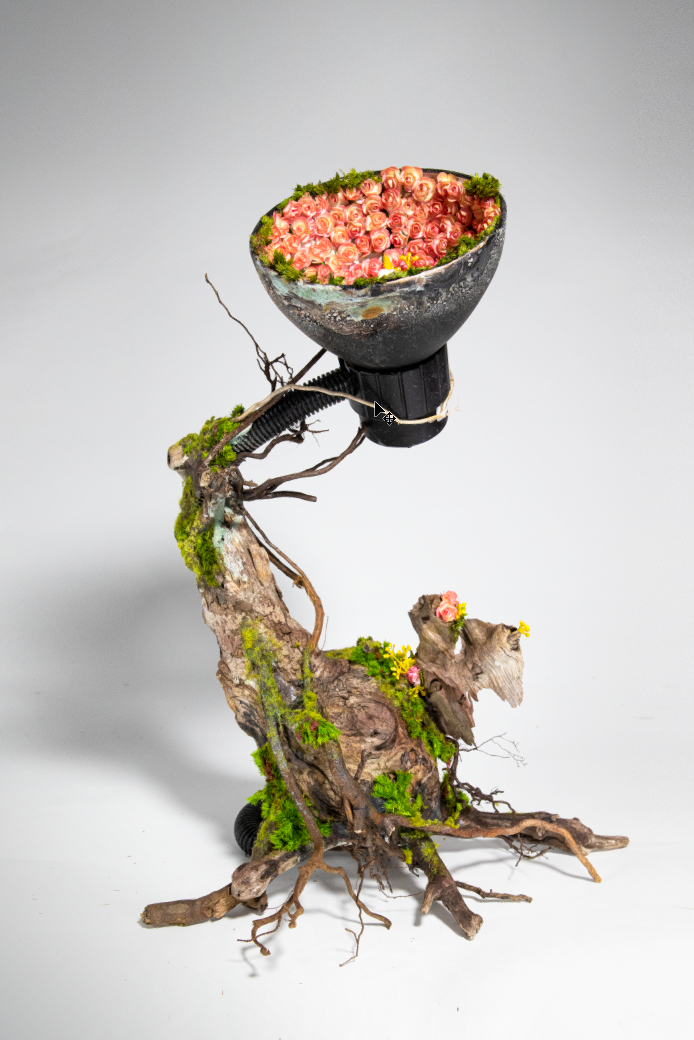 A creative flower arrangement featuring a twisted piece of driftwood with moss and small flowers, holding a black lamp shade filled with pink roses and greenery.