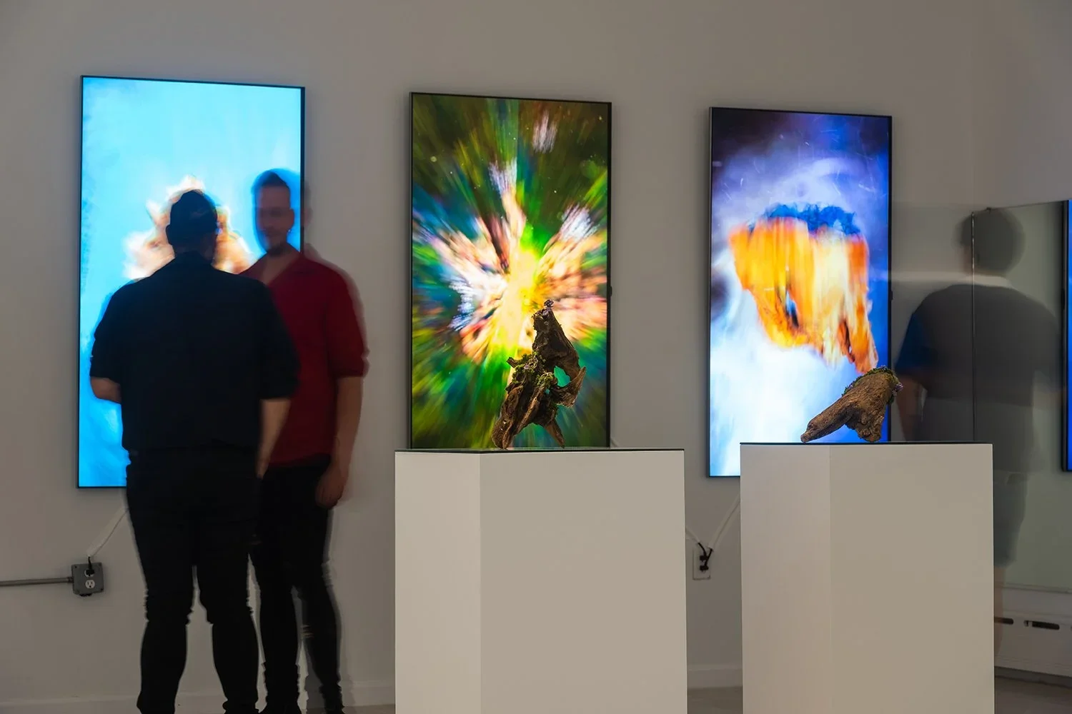 Two people are talking in front of three large digital screens displaying colorful abstract artwork. The screens are mounted on a white wall and are accompanied by sculptures on white pedestals.