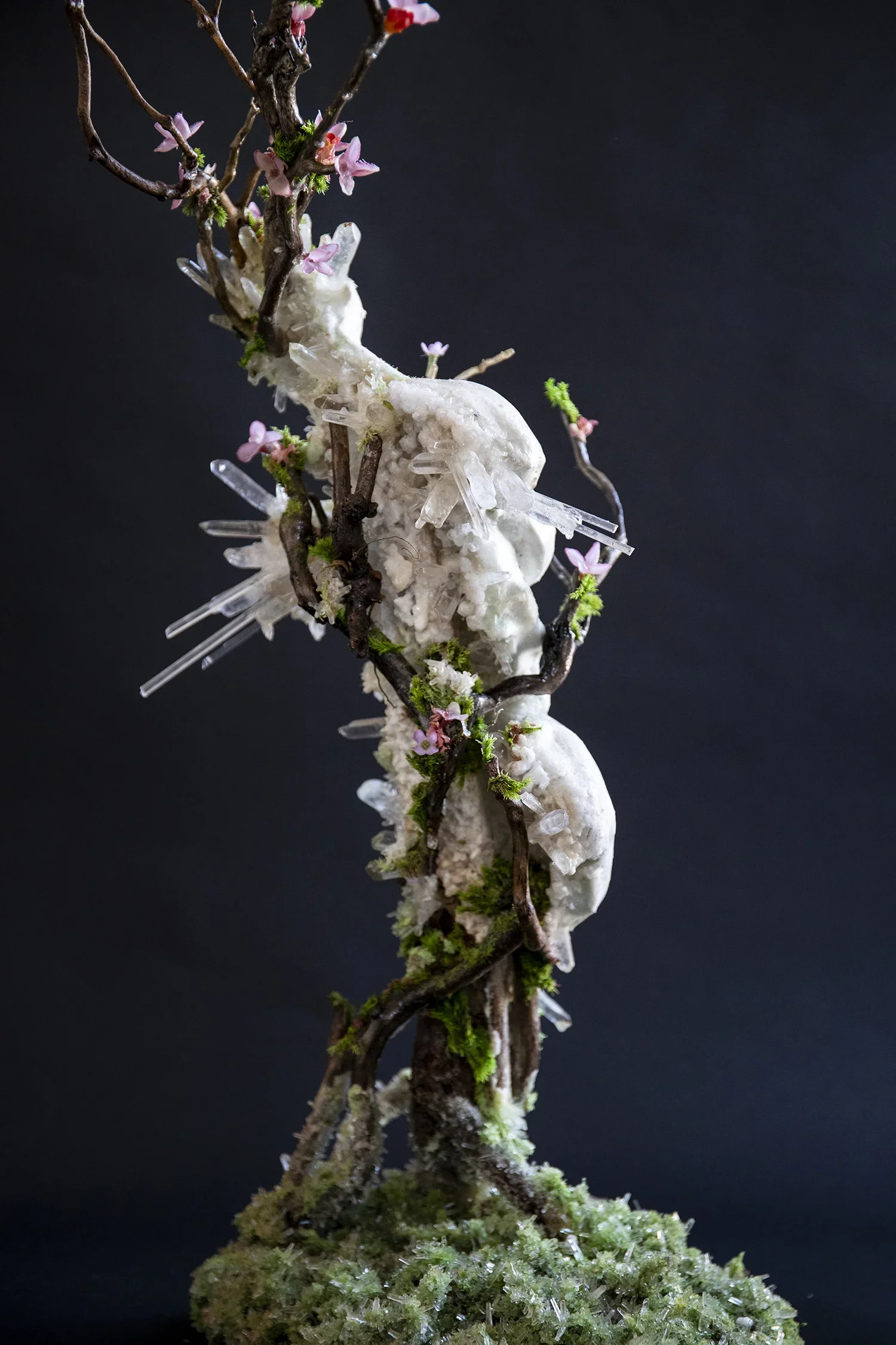 A detailed model of a tree branch with pink flowers, white foam-like material, green moss, and clear crystal formations on a black background.