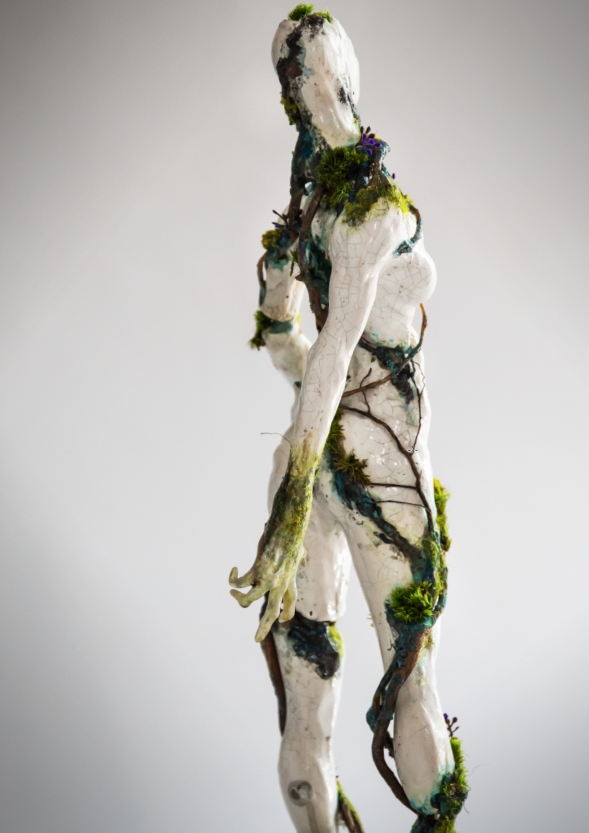 A statue of a humanoid figure with cracks and moss growing on it, resembling a female form with long arm and legs, and no facial features.