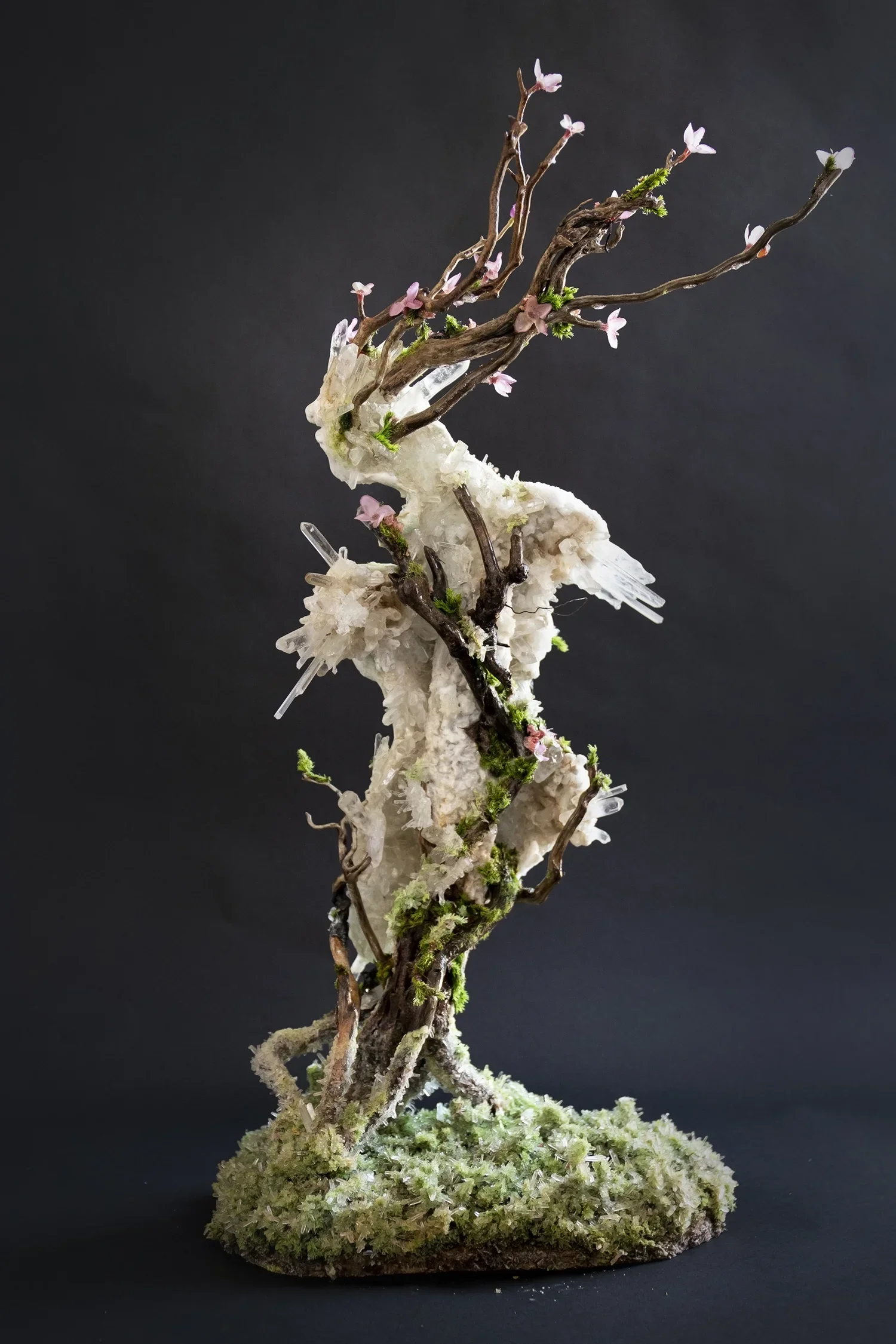 A sculpture of a tree with pink and white flowers, green moss, and some transparent acrylic elements on a black background.