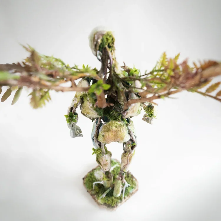 A small plant with a humanoid figure made out of moss and twigs, standing on a square base with moss, viewed from below against a white background.