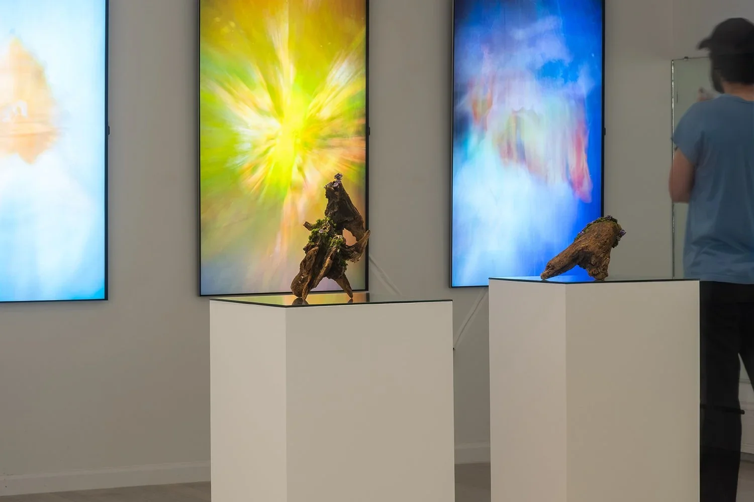 Gallery with three digital screens displaying colorful, blurred abstract backgrounds and two wooden sculptures on white pedestals.