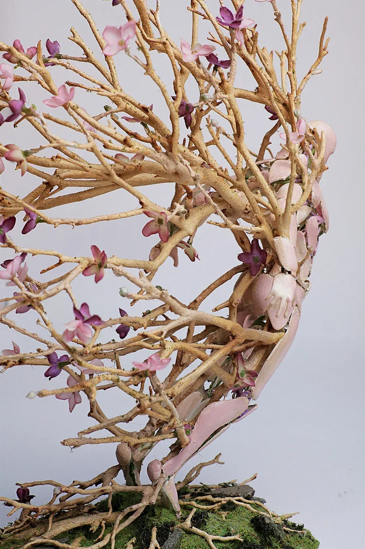 A small, pink and purple flowering tree sculpture with slender branches, placed on a moss-covered base.