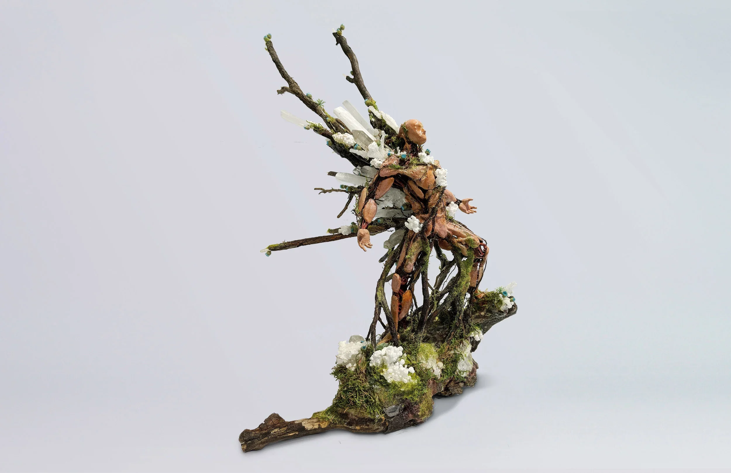 A sculpture of a fairy made from natural materials like twigs, rocks, moss, and crystals, depicted in a seated, relaxed pose with wings.