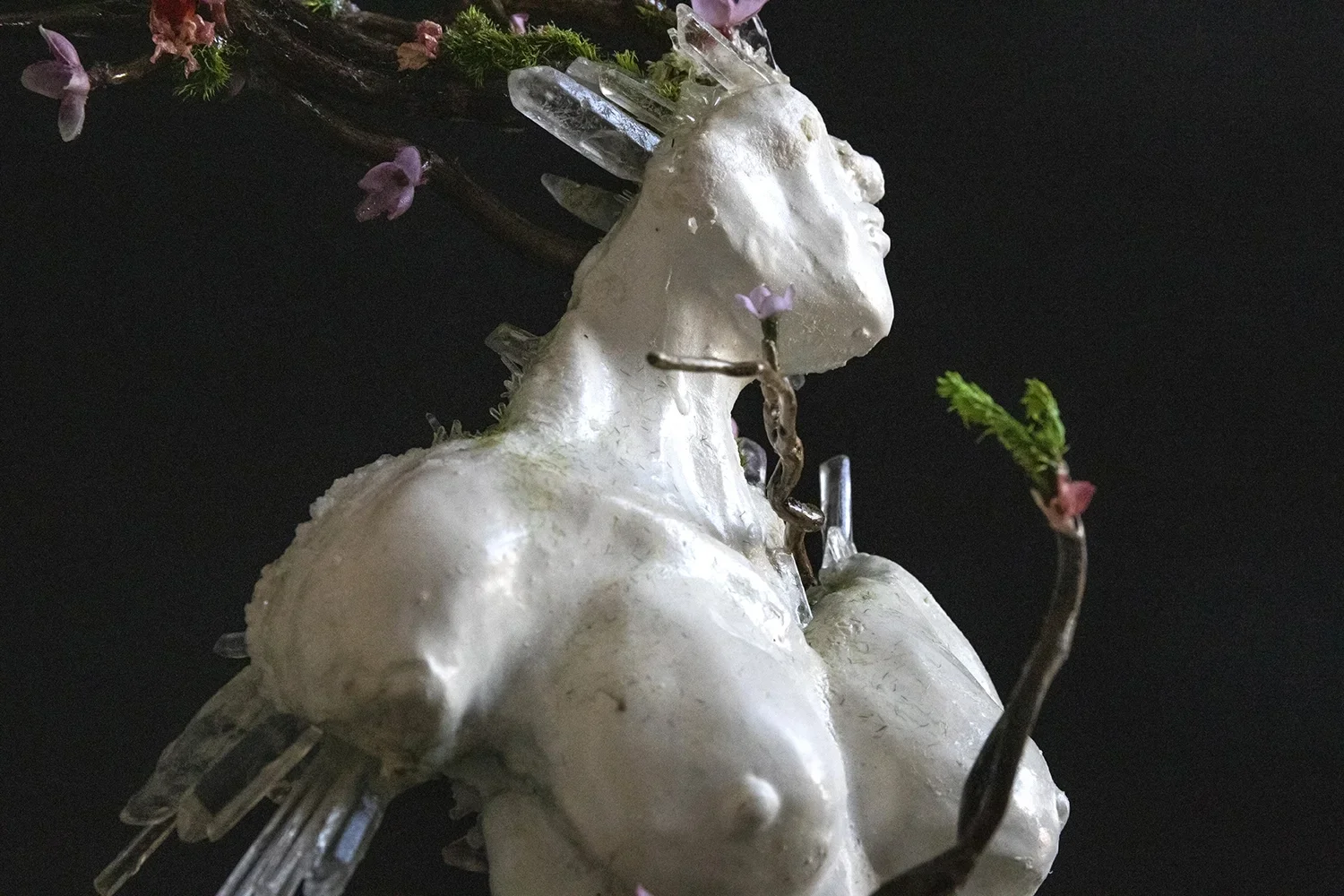 Sculpture resembling a female figure with ice crystals and small purple flowers, set against a dark background.