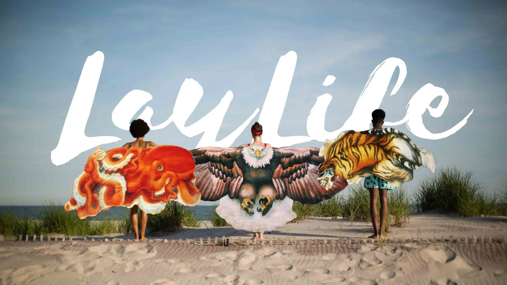 Three children holding large, colorful animal cutouts in front of a large Sign that says "Lajulie" on a beach or sandy area with grass and blue sky.