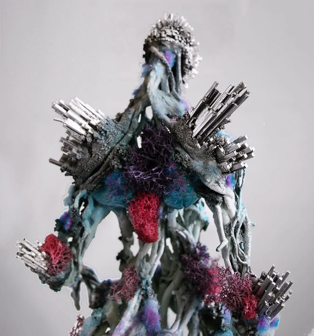 Abstract sculpture combining metallic rods, colorful textiles, and organic-looking structures resembling coral or fungi, set against a plain background.