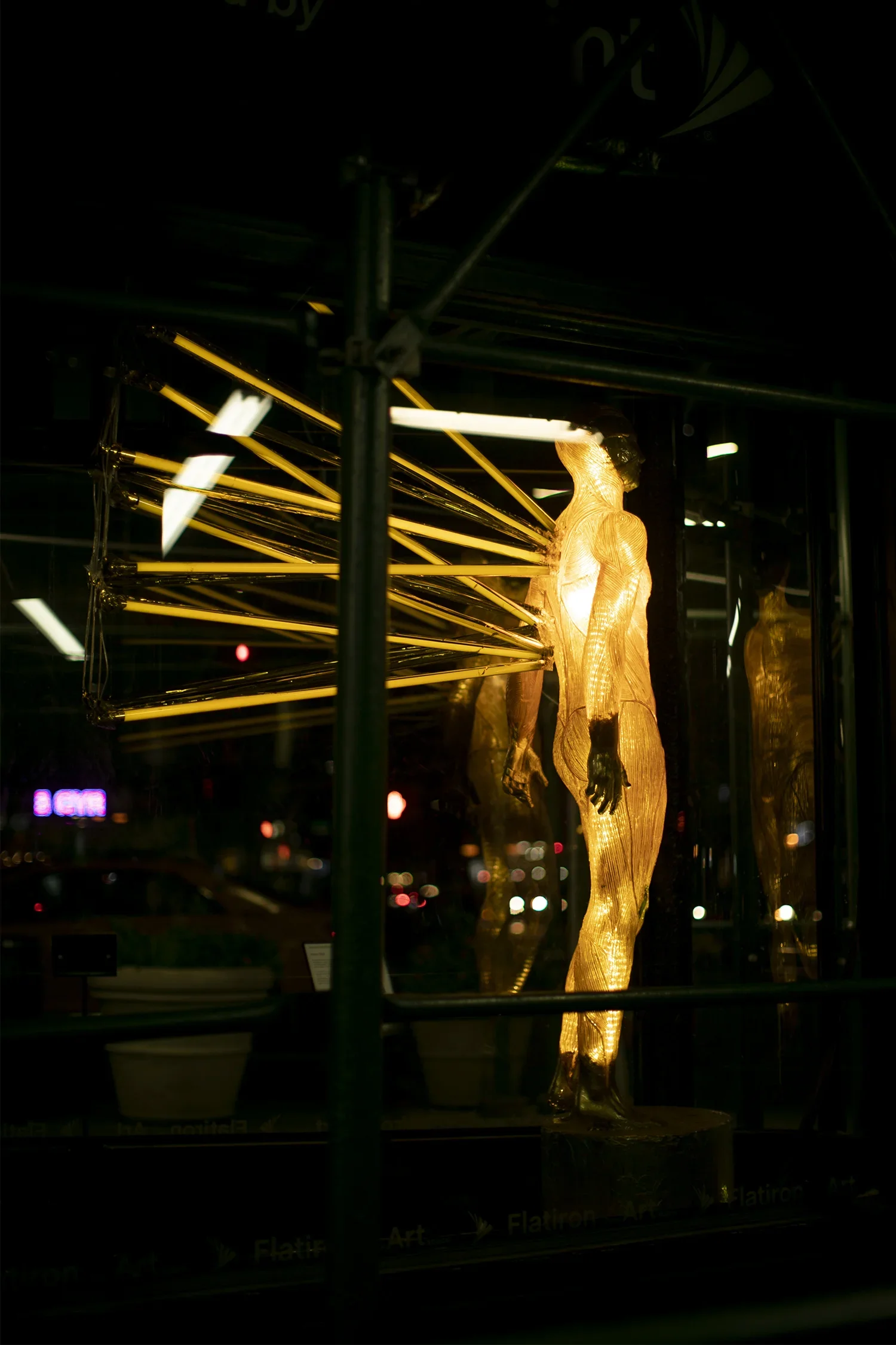 A glowing mannequin sculpture of a human figure displayed in a dark storefront window, with light reflecting off its surface.