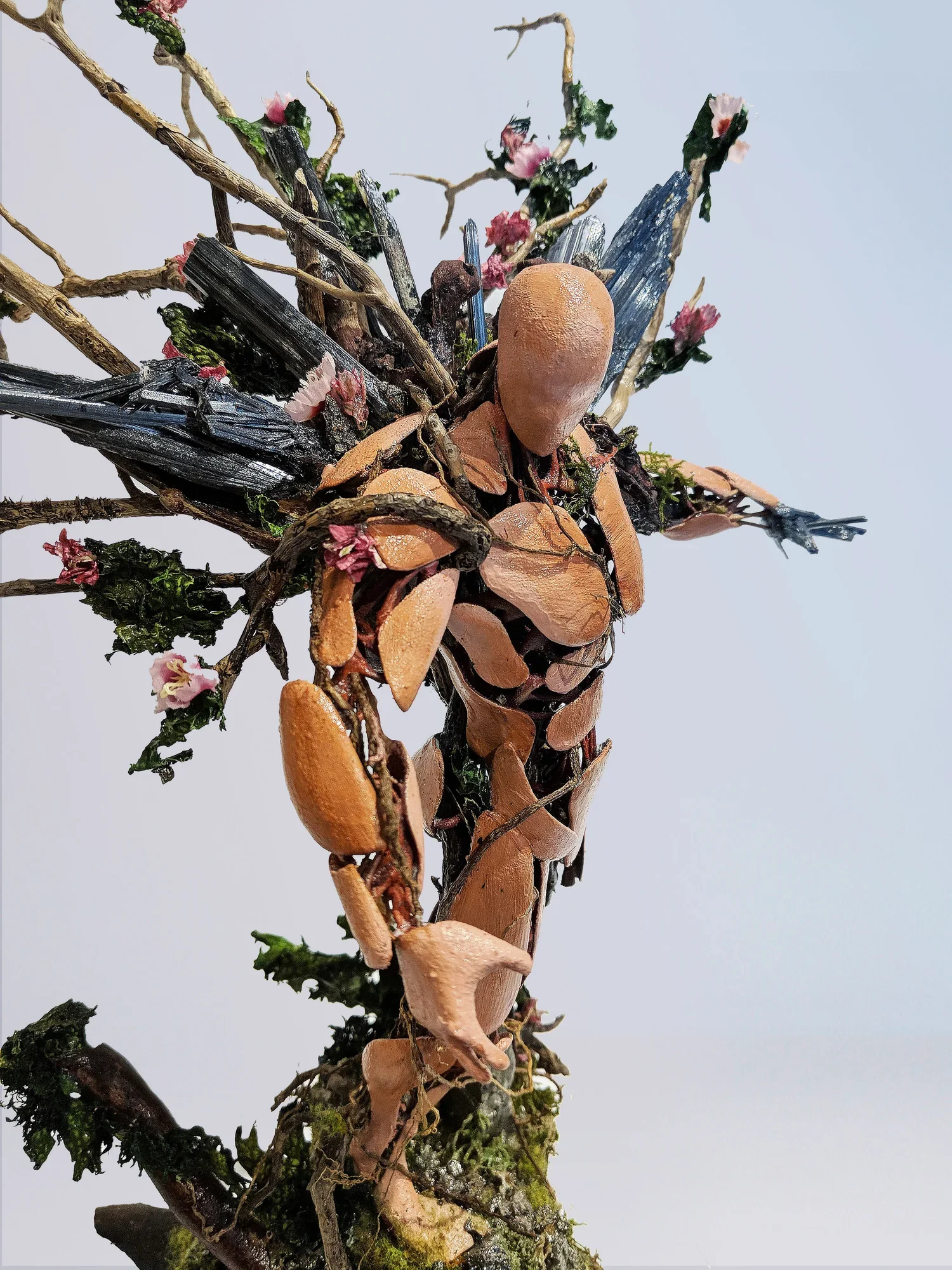 A detailed close-up of a vertically arranged natural sculpture made of twigs, dried leaves, moss, flowers, and other plant materials against a plain background.