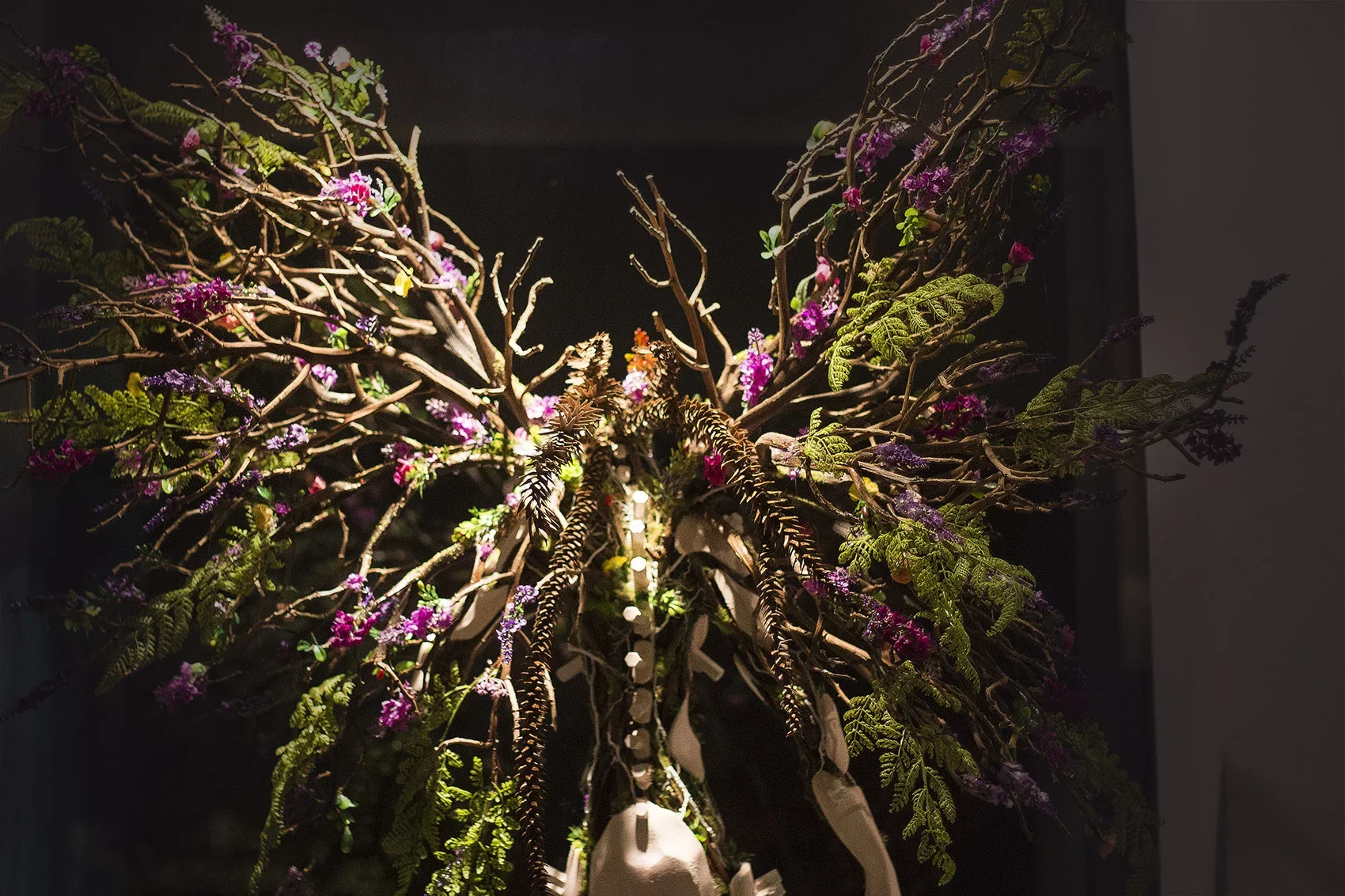 A butterfly-shaped sculpture or art installation made of intertwined branches and decorated with pink, purple, and green plants, illuminated against a dark background.